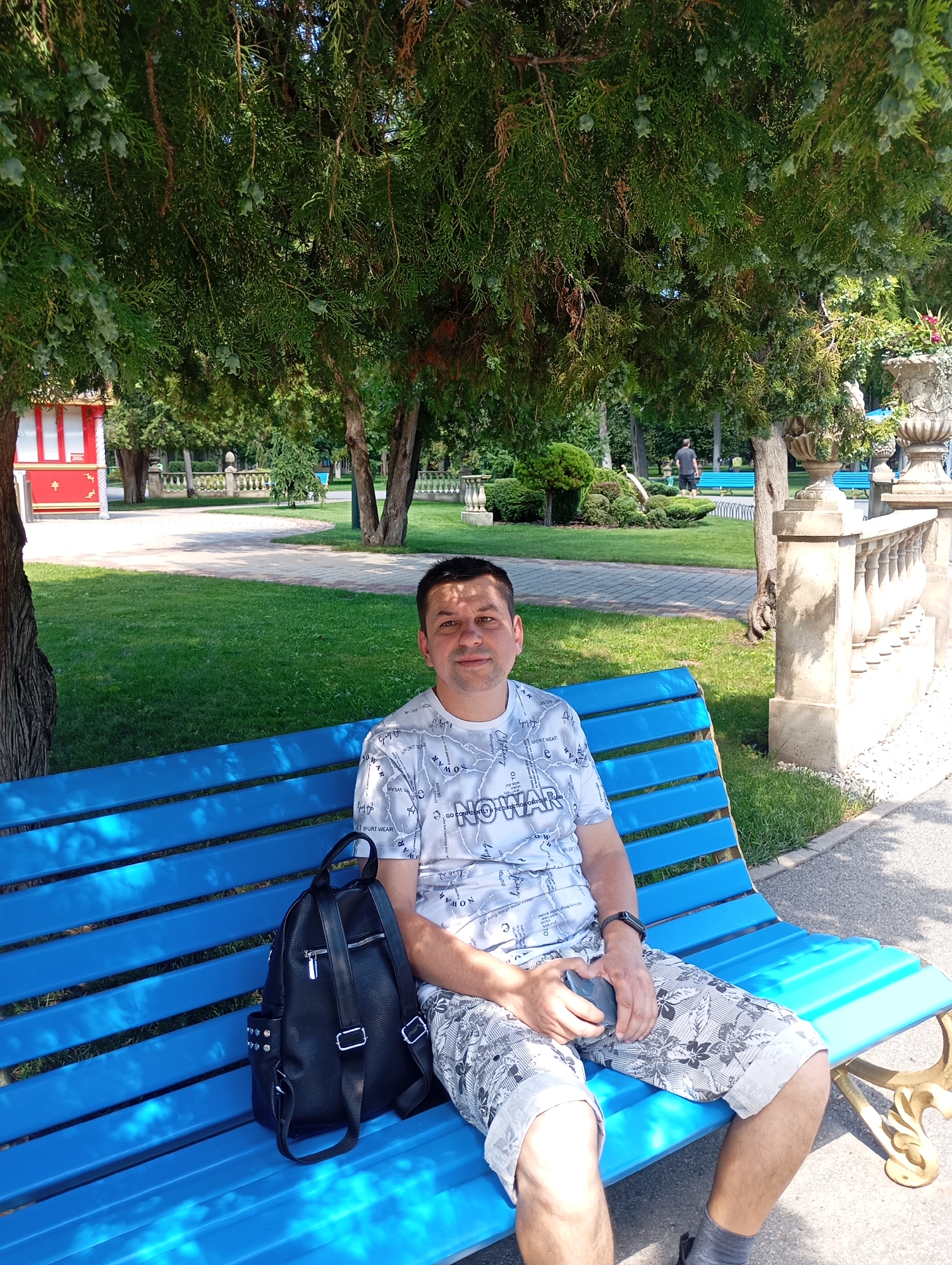 A man is sitting on a blue park bench in Kharkiv, Ukraine. He is wearing a white t-shirt with a black and white pattern and patterned shorts. A black backpack is on the bench next to him, and he is holding a grey mobile phone in his right hand. The bench is under the shade of a large tree with dense green foliage. In the background, there is a paved pathway, a manicured lawn, and ornamental stone balustrades. Further in the distance, a red and white structure resembling a kiosk or pavilion is visible, along with other park benches and trees. The lighting suggests it is a sunny day.