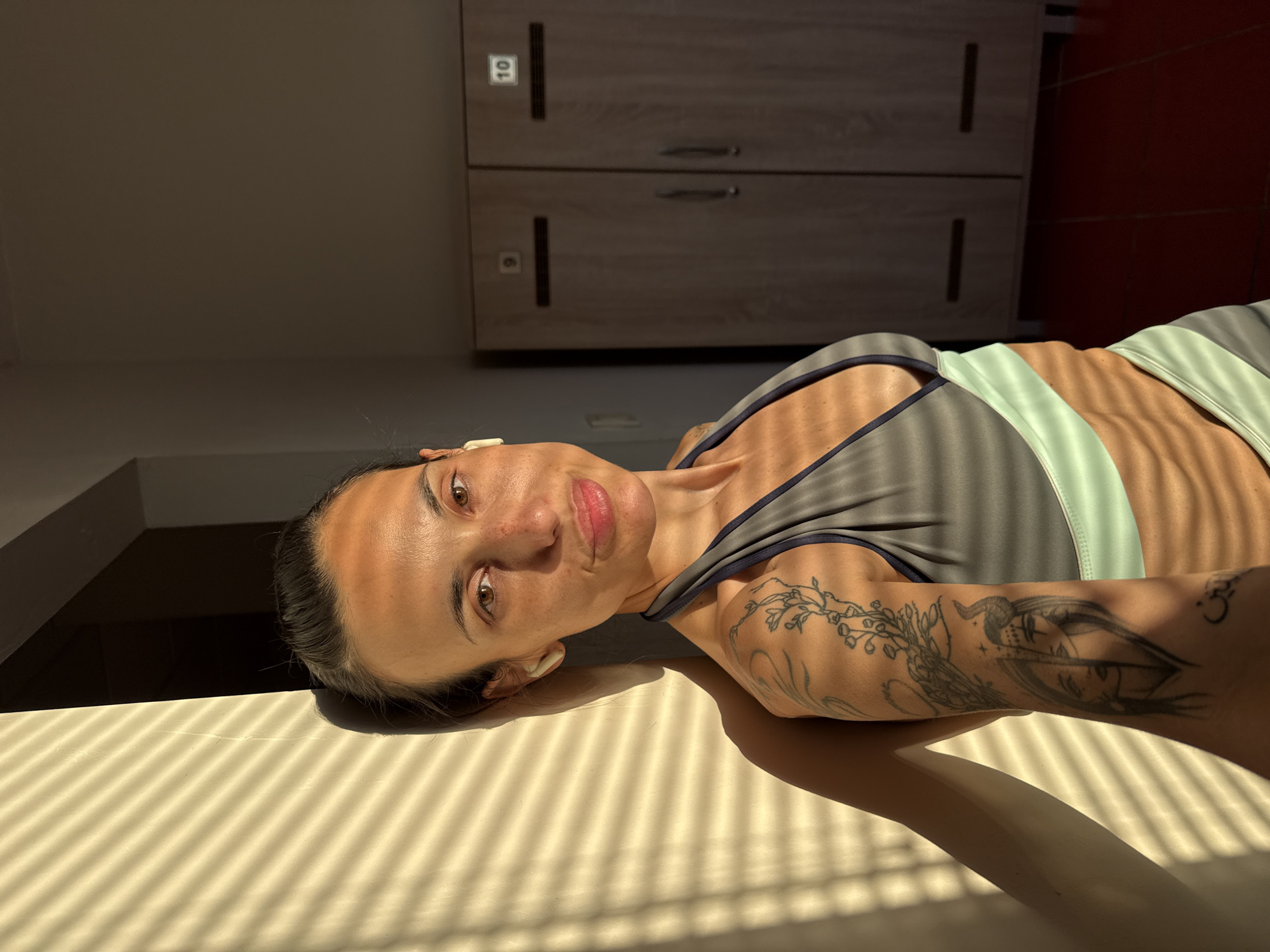 A woman lies on a light-colored surface with parallel shadows cast by window blinds. She wears a gray and light green sports bra and has a tattoo on her right arm. She has dark hair pulled back from her face, brown eyes, and wears a white wireless earbud in her left ear. In the background, there are lockers with a visible number "10" on one of them. The location is likely a changing room or a similar interior space. The location cannot be determined to be in Pleven, Bulgaria, from the given image alone.