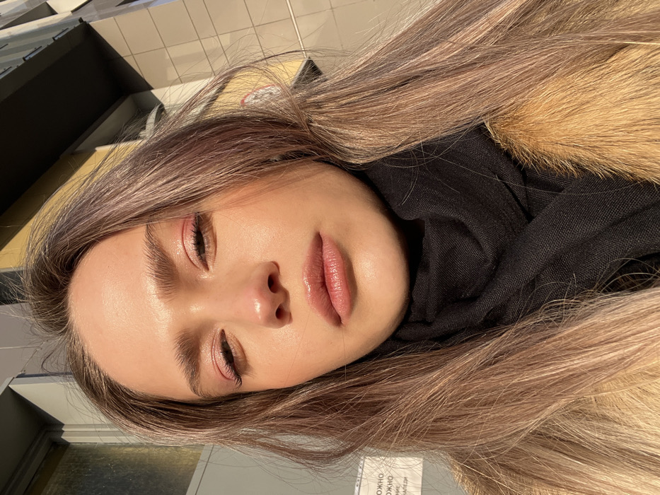 A young woman with fair skin, closed eyes, and pink eyeshadow is positioned against a blurred backdrop of buildings. She wears light lipstick and a black scarf. Her hair, a blend of blonde and light purple, cascades around her face. Golden fur trims the edge of the scarf near her hair. The lighting creates a bright, almost ethereal glow, emphasizing her features and highlighting the textures of her hair and clothing.