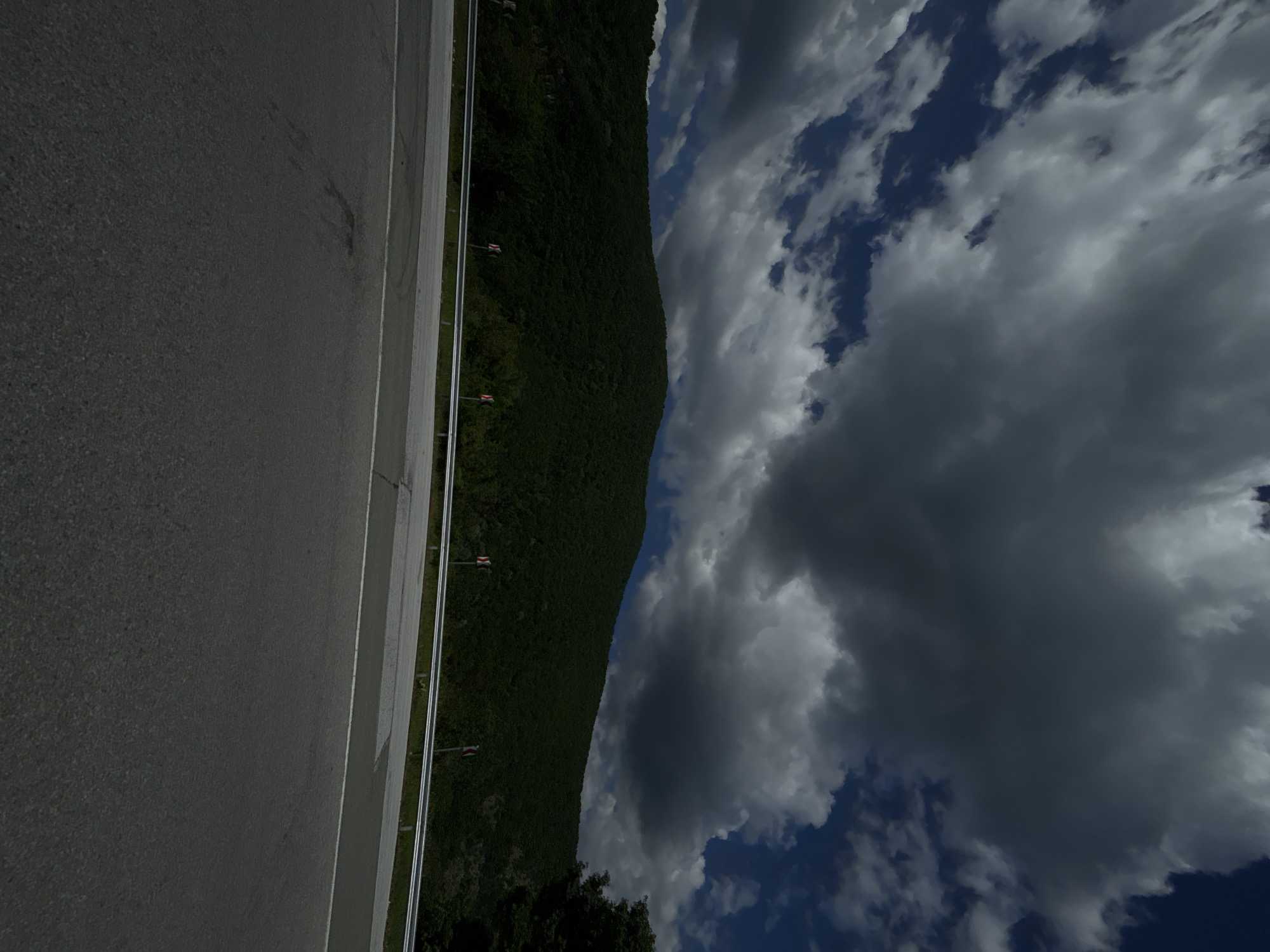 A roadside view in an unknown city shows a paved road on the left with a metal guardrail. Adjacent to the guardrail is a steep, forested hill ascending towards the sky filled with clouds. The sky is characterized by patches of blue interspersed with both light and dark storm clouds. At the bottom of the hill, some tree canopies are visible.