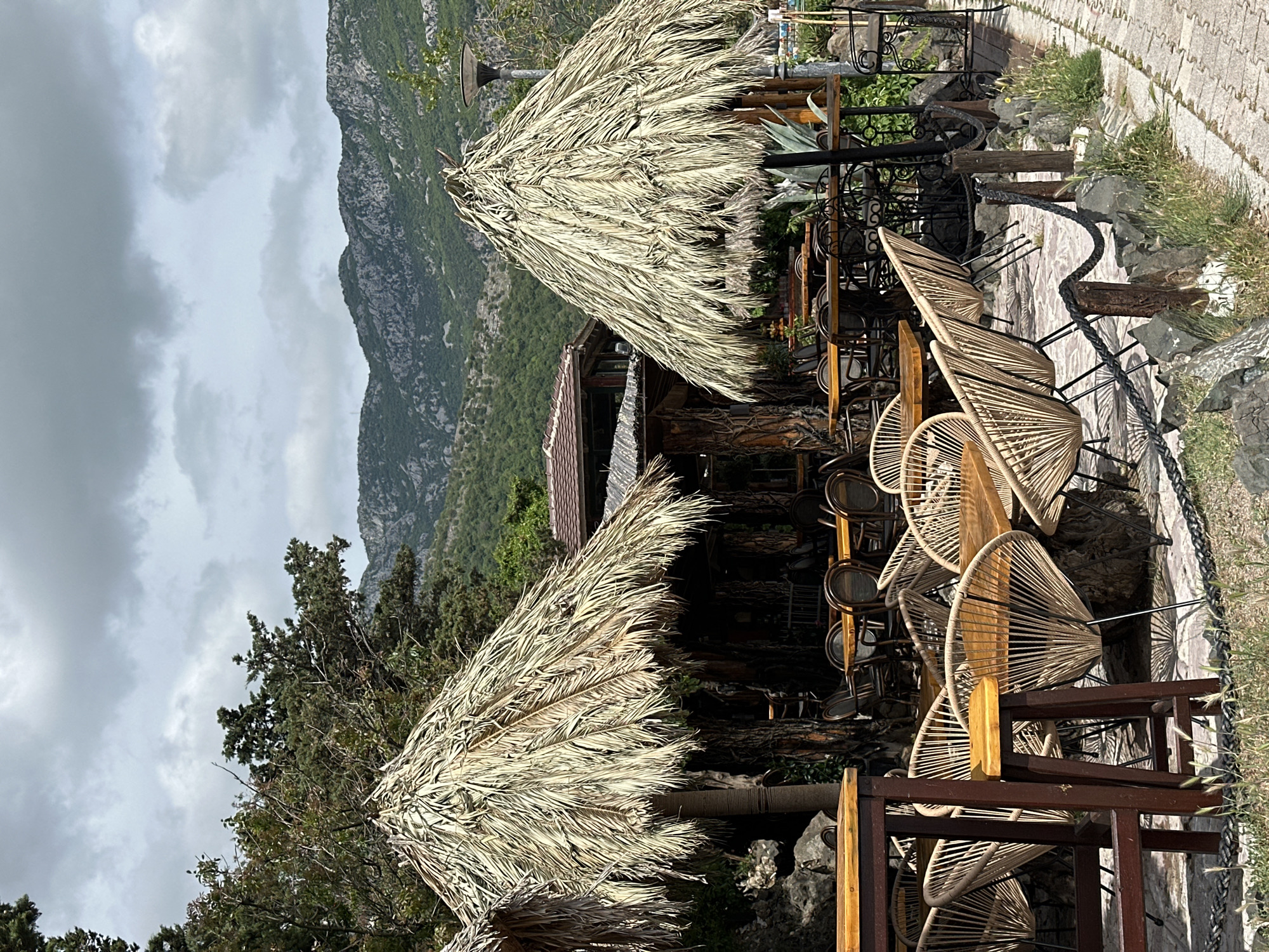 The image shows an outdoor dining area in Boreti, Montenegro. The area features tables and chairs under thatched-roof structures. In the background is a mountain range partially obscured by a cloudy sky. The tables are made of wood and paired with woven chairs. Other chairs are stacked nearby. A stone pathway is visible on the right side of the image. The overall setting suggests a restaurant or lounge with a scenic view.