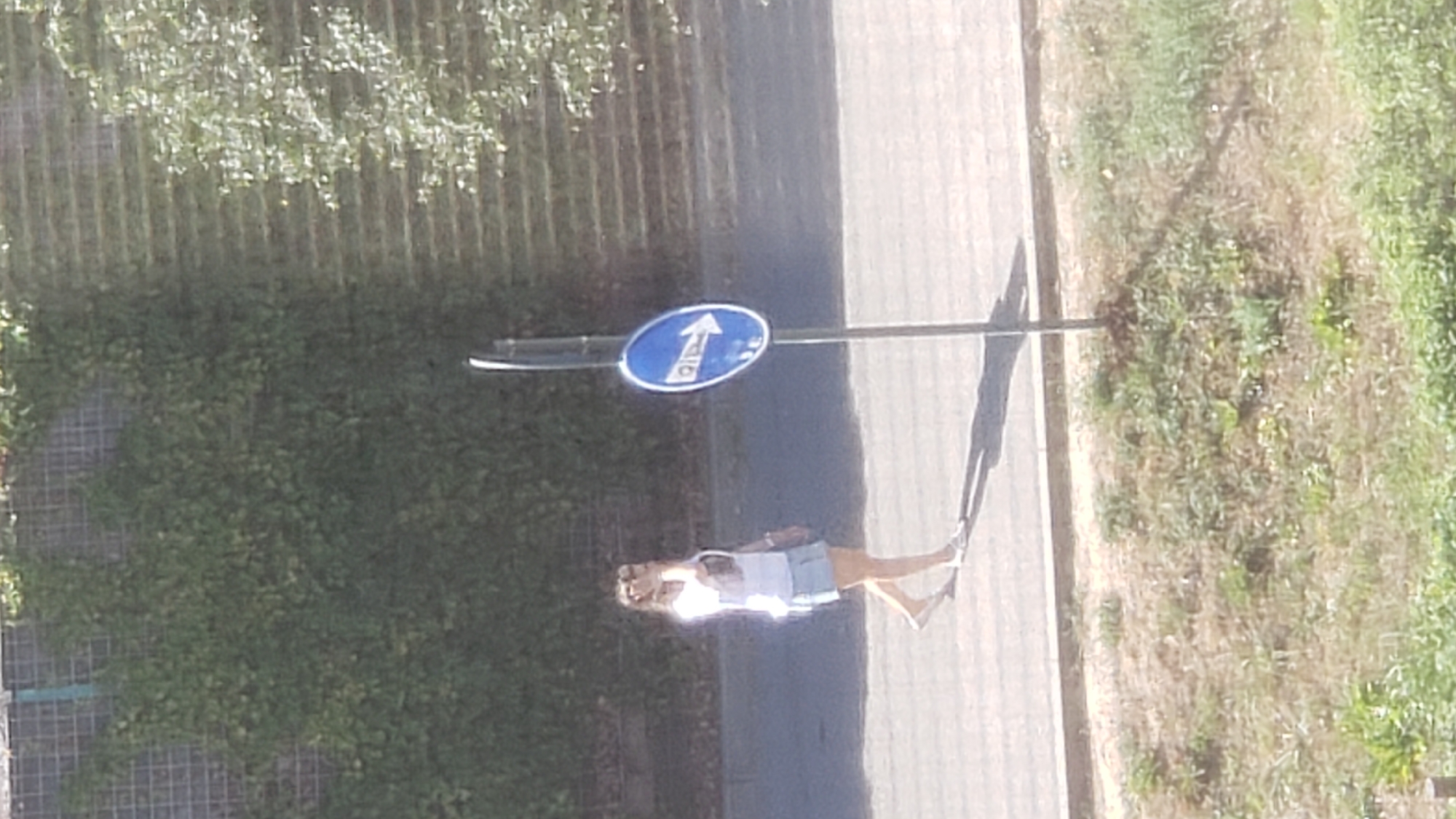 A young woman in a white t-shirt and denim shorts walks on a paved surface beside a grassy embankment. She is looking to her right. Next to her, a blue circular sign with a white arrow pointing to the right is mounted on a metal pole. The background consists of a textured wall covered in greenery, and a further section of foliage. The scene appears to be an outdoor urban environment. The specific location in Prešov, Slovakia, cannot be determined from the image alone.