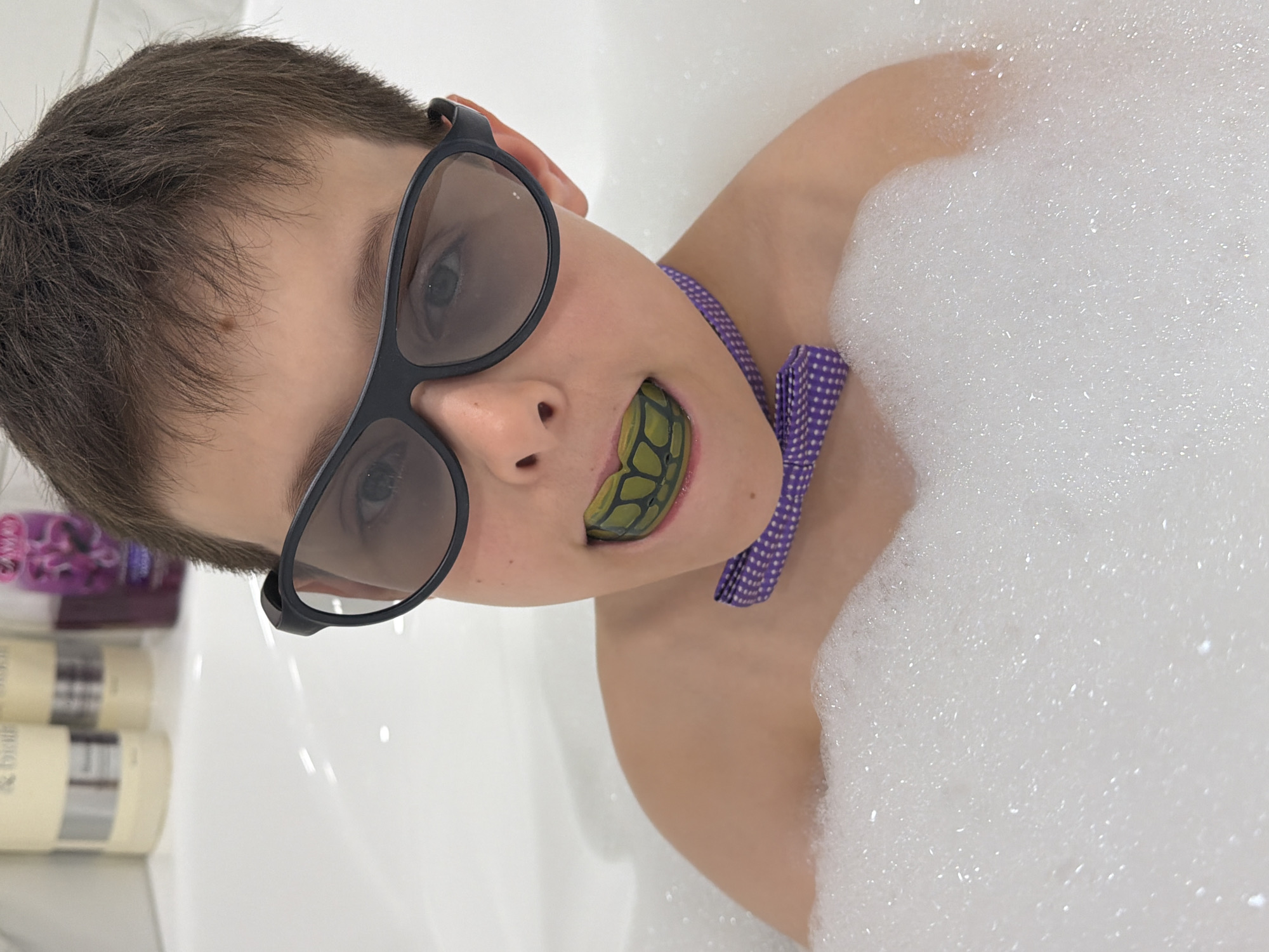 A close-up, medium-shot photograph shows a young person in a bathtub filled with bubbles. The person is wearing black-rimmed glasses and a purple polka-dot bow tie. The person also has a green and black mouthguard in their mouth. There are bottles of bath products visible on the edge of the bathtub. The location is unknown and there is no indication in the image that the location is in the city of Votkinsk, Russia.