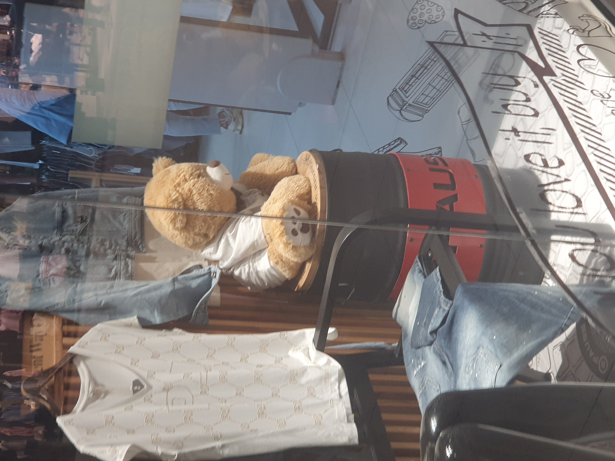The image shows a retail display, likely inside a store window in Plovdiv, Bulgaria. A large, light-brown teddy bear reclines inside a black and red cylindrical container, possibly a promotional item. Clothing is also visible: a white patterned t-shirt on a hanger, as well as denim jeans. The background features a reflected cityscape, and a white surface with hand-drawn elements. The writing on the glass includes the words "Love it...Buy".