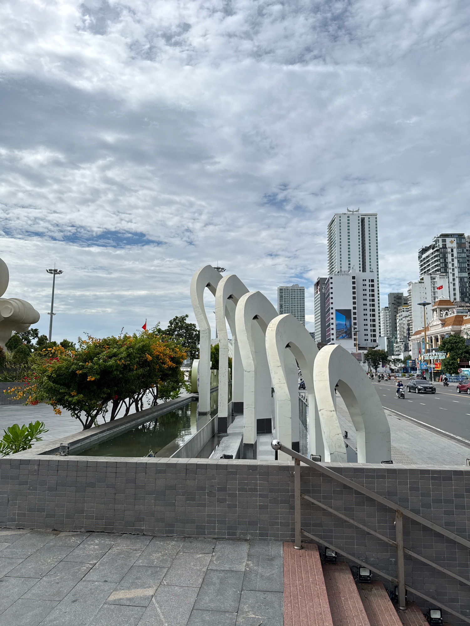 This is a street-level, eye-level, wide shot of the Nha Trang coastline in Vietnam on a cloudy day. In the foreground, a dark grey tiled wall runs horizontally across the image, with a set of red-brown stairs to the right leading upwards. A silver railing follows the stairs. Beyond the wall is a long, narrow water feature. To the left of the water feature is a large, abstract white sculpture with multiple arched elements. Further left, a large white statue is partially visible.
Behind the water feature and sculpture, a line of trees with green leaves and bright orange and red flowers runs along the edge of the water. A red flag is visible on a pole amidst the trees. In the background, a multi-lane road runs parallel to the coastline, with traffic including cars and motorcycles. The road is lined with modern high-rise buildings of varying heights and architectural styles. One prominent building is a white skyscraper with green accents, identified by a "NOVOTEL" sign. Other buildings are also visible, some with balconies and windows.
The sky is filled with dramatic, layered grey clouds, with patches of lighter blue sky visible in places. Streetlights are visible along the road, and the overall environment suggests a public space with urban development and natural elements. The scene is static, with no obvious interactions between people or subjects captured at this moment. The lighting is diffused due to the cloud cover, creating soft shadows.