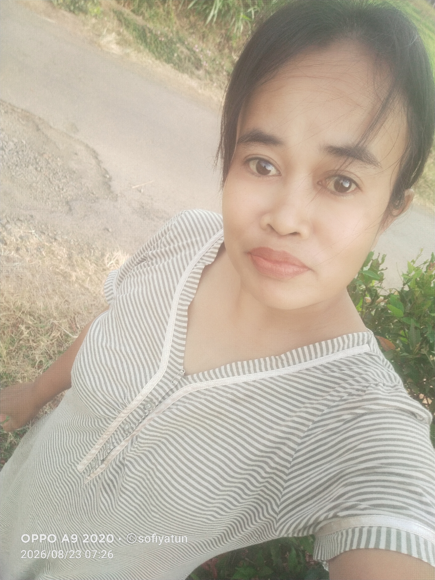 A woman is taking a selfie outdoors, likely in the city of Cluwak, Indonesia, based on the overlaid text which reads "OPPO A9 2020 - ©sofiyatun 2026/08/23 07:26". She is wearing a grey and white striped dress and her hair is pulled back. The background shows a dirt road, dry grass, and some greenery. The lighting suggests it is daytime.