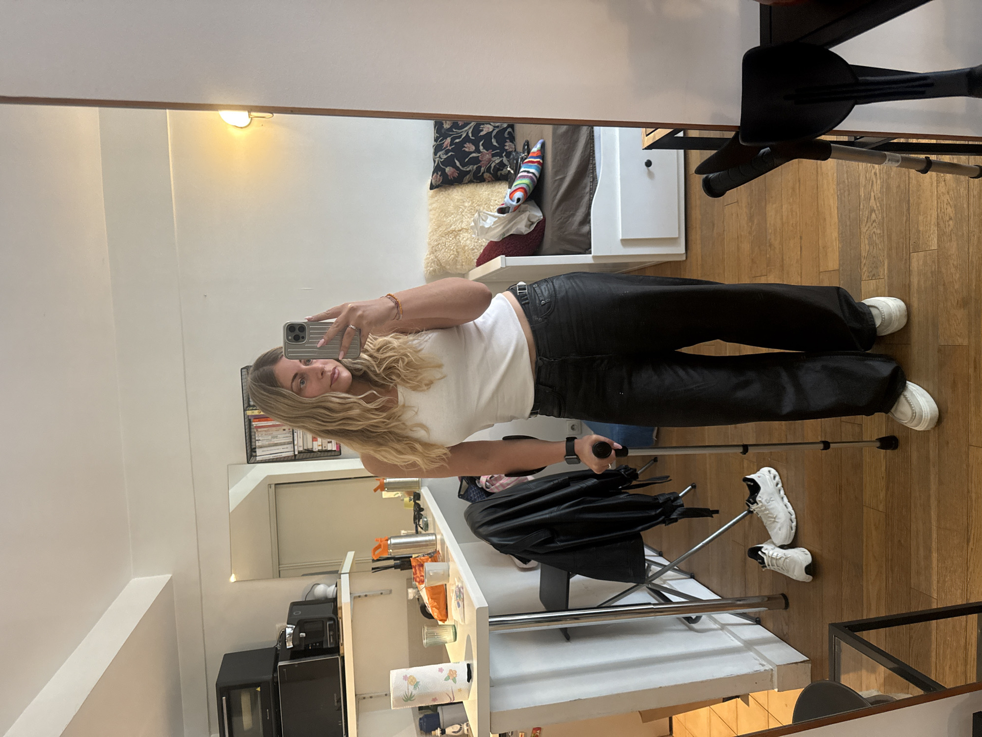 A young woman with long, wavy blonde hair takes a mirror selfie in what appears to be a compact apartment. She is dressed in a white sleeveless crop top, black high-waisted pants, and white sneakers. A black belt cinches her waist, and she wears a watch on her left wrist, rings on her left hand, and bracelets on her right wrist. Her nails are painted light pink. She holds a silver smartphone in her left hand, capturing her reflection, and is holding a single crutch with her right hand, suggesting a mobility aid. Another pair of white sneakers rests on the light wooden floorboards next to a second pair of crutches.
The mirror reflects the interior of the apartment, showcasing a combined living space. To the woman's right, a bed is visible with dark bedding, a patterned pillow, a fluffy beige throw, and a colorful striped cushion. A white nightstand with a lamp stands beside the bed. To her left, a white counter or shelf unit holds various items, including a paper towel roll, glass cups, metallic bottles, and small black kitchen appliances such as a microwave and another device. A black metal rack filled with books is mounted on the wall above this area. A black jacket is draped over a nearby chair or stand.
The apartment has light-colored walls and warm wooden flooring, suggesting a modern and functional design. A warm, illuminated light fixture is visible on the wall above the bed area. The scene depicts a casual moment in a domestic setting during the daytime, possibly in Paris, indicating a temporary or small urban dwelling. No clear text is legible in the image.