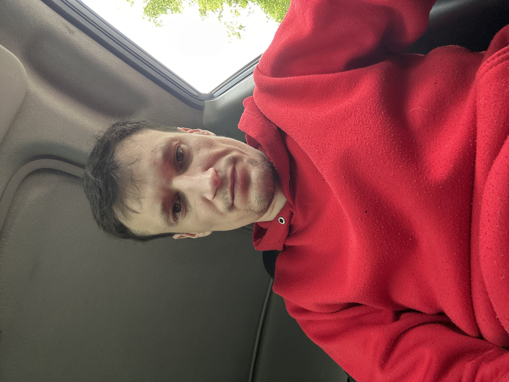 A close-up, low-angle, indoor selfie shows a light-skinned male with short, dark hair, and a neutral expression. He is wearing a red hooded sweatshirt with the hood partially up. The background consists of the interior of a vehicle, with the headliner and part of the window visible. Outside the window, trees and sky can be seen. The image appears to be taken in Makiv, Ukraine.