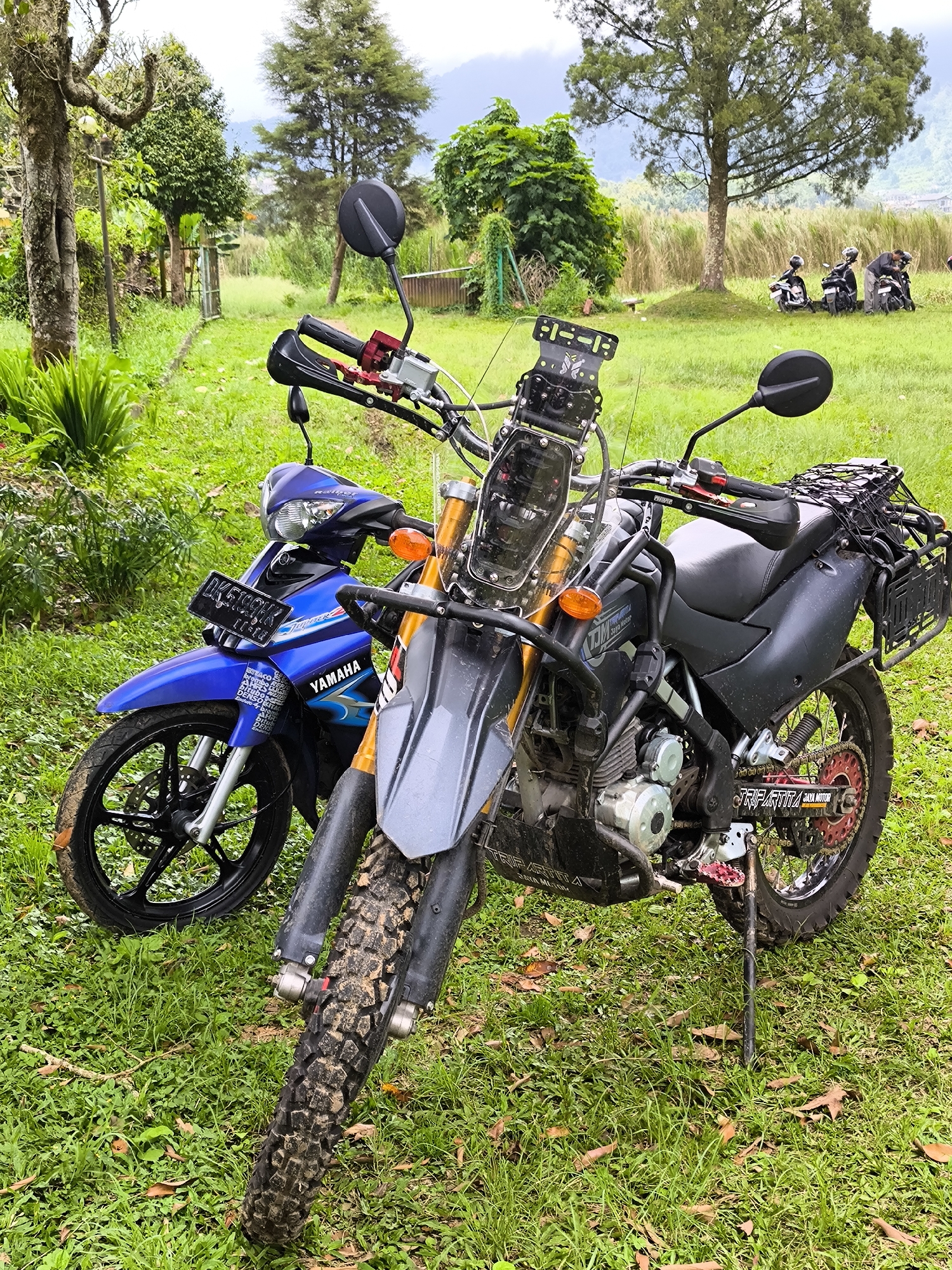 The scene depicts two motorcycles parked on a grassy field in Bedugul, Indonesia. The first motorcycle is a blue Yamaha, with license plate DK 61CQH, and is situated to the left of a larger, grey dirt bike. The dirt bike has a windshield and several attachments, and appears to have been driven off-road, indicated by the mud on its tires.
In the background, three individuals are standing near additional motorcycles. The environment includes lush green grass, trees, and a slightly blurred mountainous landscape in the distance. The scene is captured in daylight.