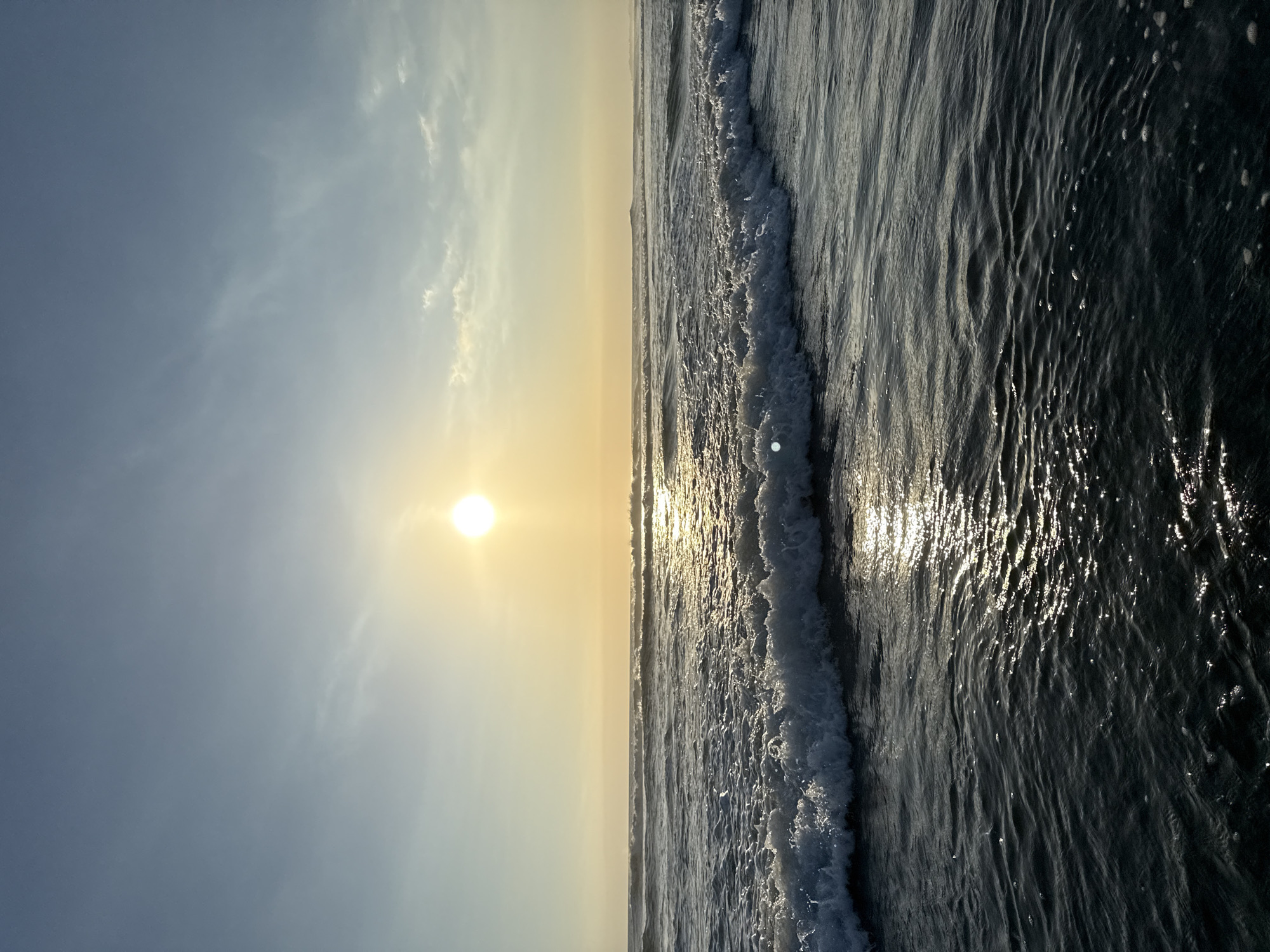 The image depicts a serene ocean scene with waves crashing on the shore under a pale yellow sky, suggestive of either sunrise or sunset. The sun, a bright orb, is positioned high in the sky, casting a warm glow and reflecting shimmering patterns on the water's surface. The ocean is a deep, dark blue, with white foam cresting the waves. The sky is a gradient of pale yellow near the horizon, transitioning to a soft blue at the top, with a few wispy clouds scattered across it. The scene is devoid of any human presence or man-made structures, presenting a natural, undisturbed seascape. While the specific location in North Kuta, Indonesia, is not visually identifiable from the image itself, the general description points to a coastal environment.