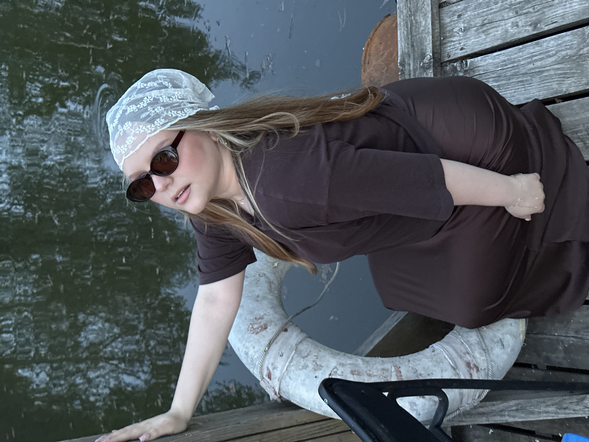 A woman is on a dock. She is wearing sunglasses and a white lace headscarf. She is wearing a brown dress. Her right hand is on the dock. A life preserver is near her. There is water next to the dock. The dock is made of wooden planks. The location is likely in Chaikovsky, Russia, as indicated by the prompt.