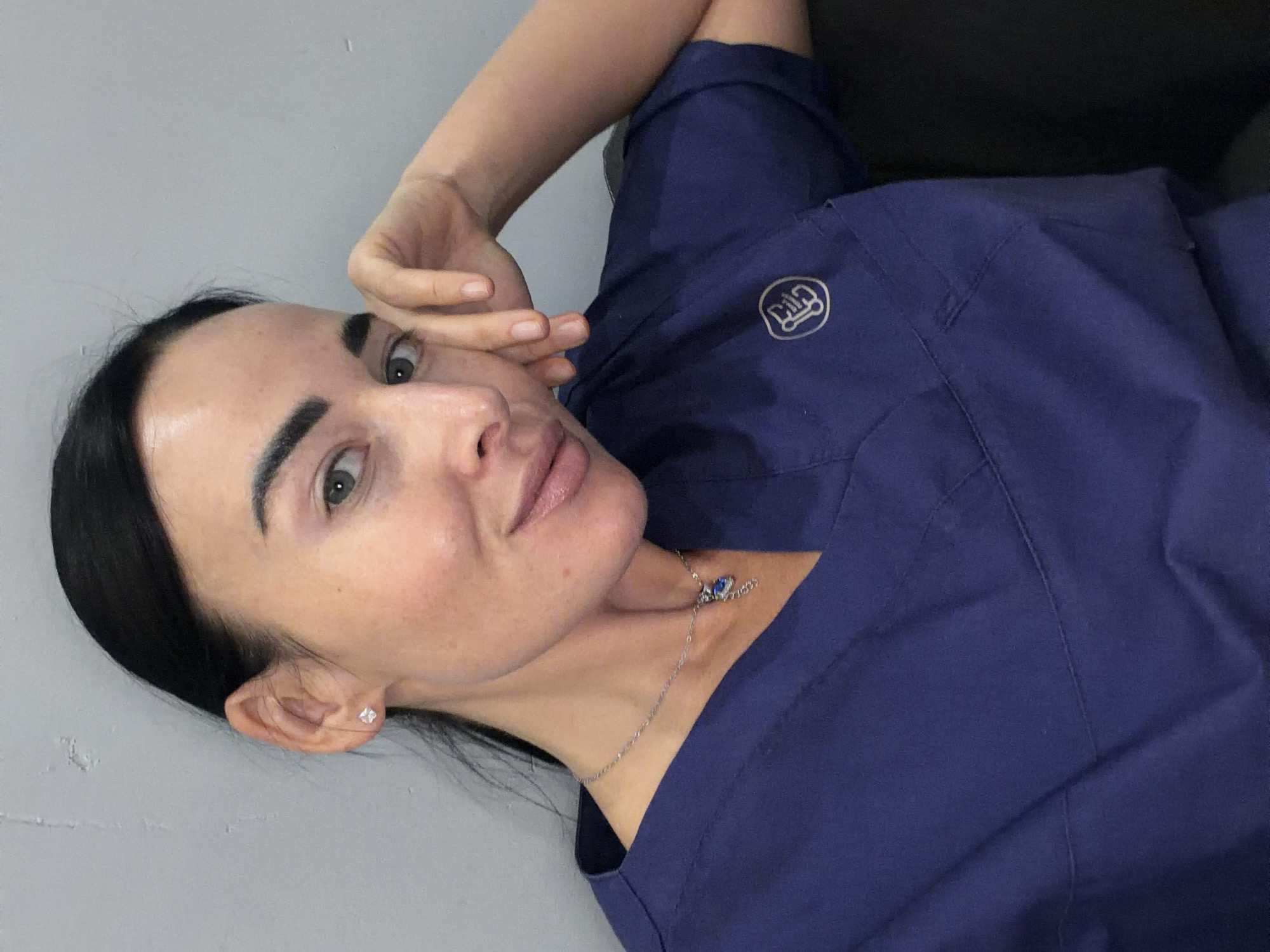 A medium close-up, angled portrait captures a woman with dark, pulled-back hair and light-colored eyes, looking directly at the camera with a neutral expression. The setting appears to be indoors, against a plain light grey wall, with a darker, indistinct surface behind her right shoulder. This scene is located in Saint Petersburg, Russia.
The woman is wearing a dark blue scrub top, which has a circular white logo on the upper left chest area. The logo features stylized letters, possibly "Gia" or "GH". Around her neck, she wears a delicate silver-colored chain necklace with a small, blue, butterfly-shaped pendant. A small, clear or silver stud earring is visible in her left earlobe. Her right hand is raised, with fingers lightly touching the side of her face near her temple. The image is a posed shot, focusing on her upper body and face.