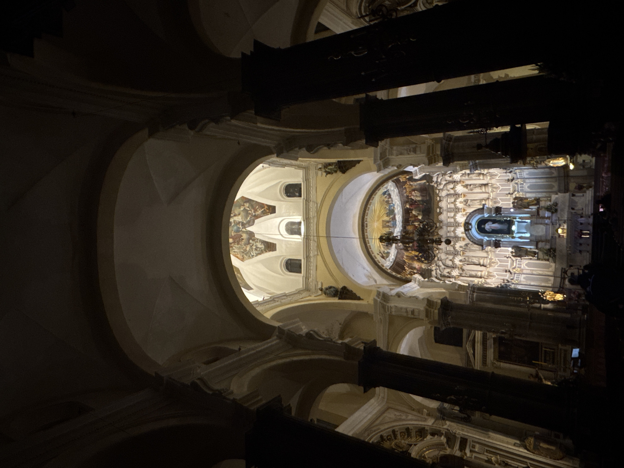 The image shows the interior of a baroque-style cathedral in Puebla, Mexico. The view is looking upwards, with the main altar visible in the distance. The cathedral's architecture features a series of arches and ornate columns. Frescoes adorn the ceilings. The scene is dimly lit, with light focused on the altar and surrounding areas. There are no people visible in the image.