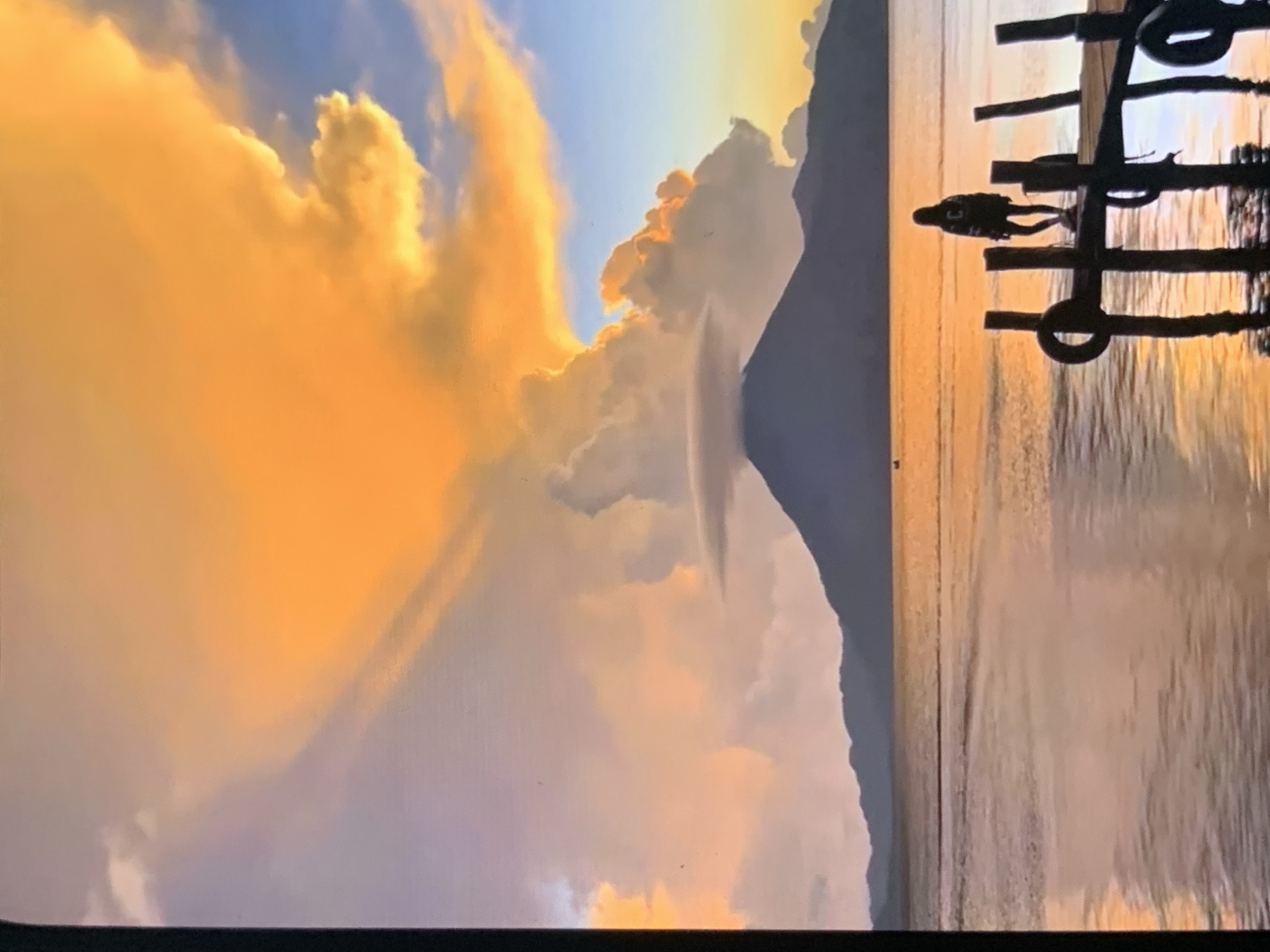 This media file captures a dramatic sunset over a calm body of water, likely a lake or sea. The sky is filled with vibrant orange and yellow clouds, illuminated by the setting sun. A dark, silhouetted mountain range forms a backdrop on the left side of the frame, contrasting with the bright sky. On the right, a wooden pier or jetty extends into the water, also in silhouette. A small, dark figure of a person is visible on the pier, appearing to be standing or walking. The water reflects the warm colors of the sky, creating a serene and picturesque scene. The overall impression is one of natural beauty and tranquility. The time of day is clearly sunset, given the intense colors in the sky and the low angle of the sun. The weather appears clear, allowing for an unobstructed view of the celestial display. While specific location cues are limited, the presence of a mountain range and a pier suggests a natural landscape with some human development. No visible text is present in the image.
