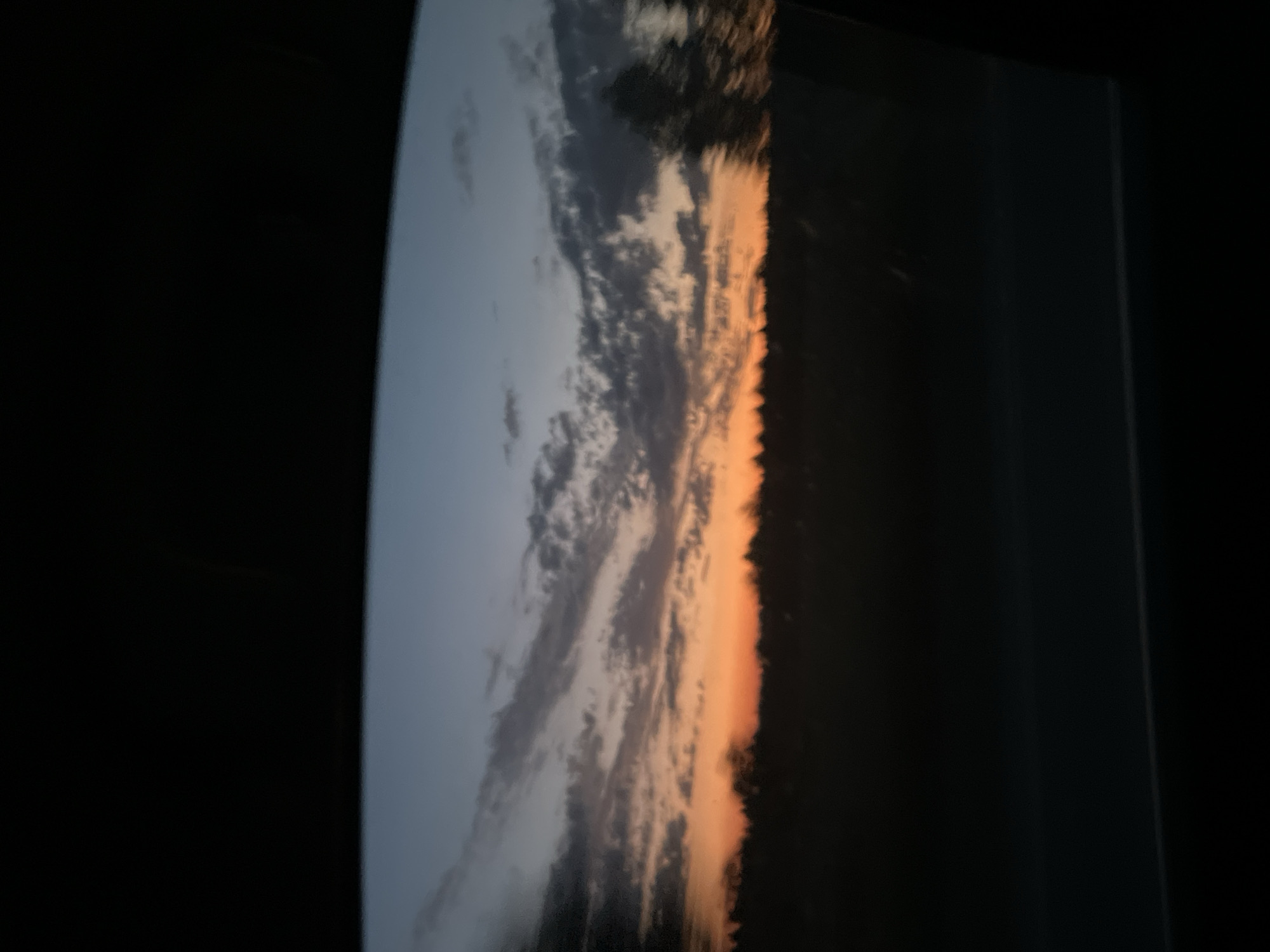The image depicts a dusky sky viewed through a car window, with a dark silhouette of trees or a treeline in the distance. The sky exhibits a gradient of colors, transitioning from a deep orange near the horizon to lighter shades of blue and gray higher up. Wispy clouds are scattered across the sky. The foreground is dominated by the dark interior of the car, suggesting the photo was taken while in motion. There are no people or distinct objects visible, only the natural landscape and the car's interior framing the view. The setting appears to be rural or on the outskirts of a settlement, potentially in or near the village of Поварёнки, Russia, given the description.