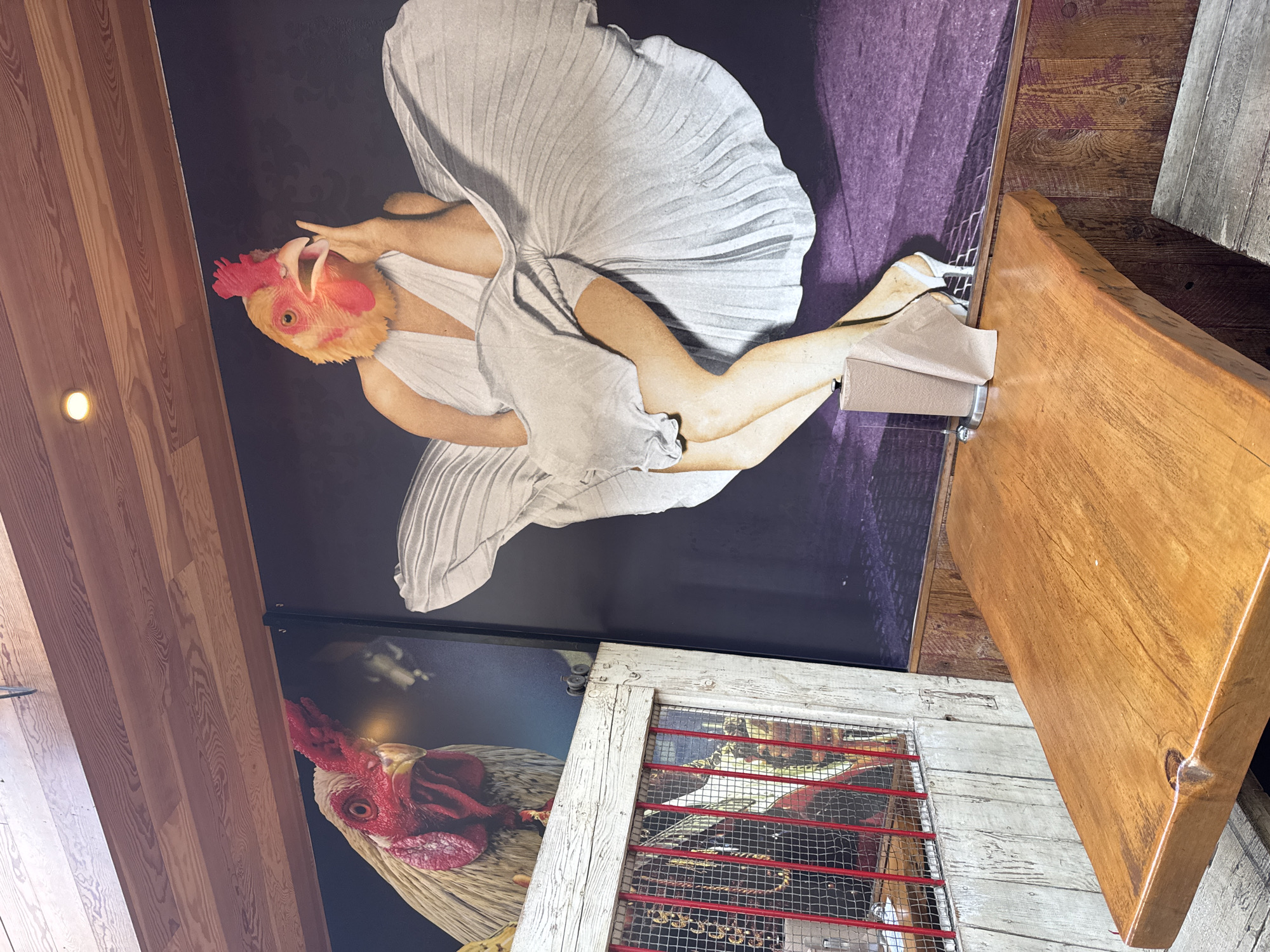 This image shows the interior of a space in Costa Mesa, United States of America, featuring eclectic decorations. On the wall are large posters: one of a woman in a Marilyn Monroe-esque pose, but with a chicken head, and another with a close-up of a rooster. A decorative repurposed wooden object, potentially an old door or window frame, with red-painted bars and metal mesh, is also on the wall. Part of a wooden countertop or table is visible, suggesting a commercial establishment like a restaurant or bar. There are no people visible in the scene.