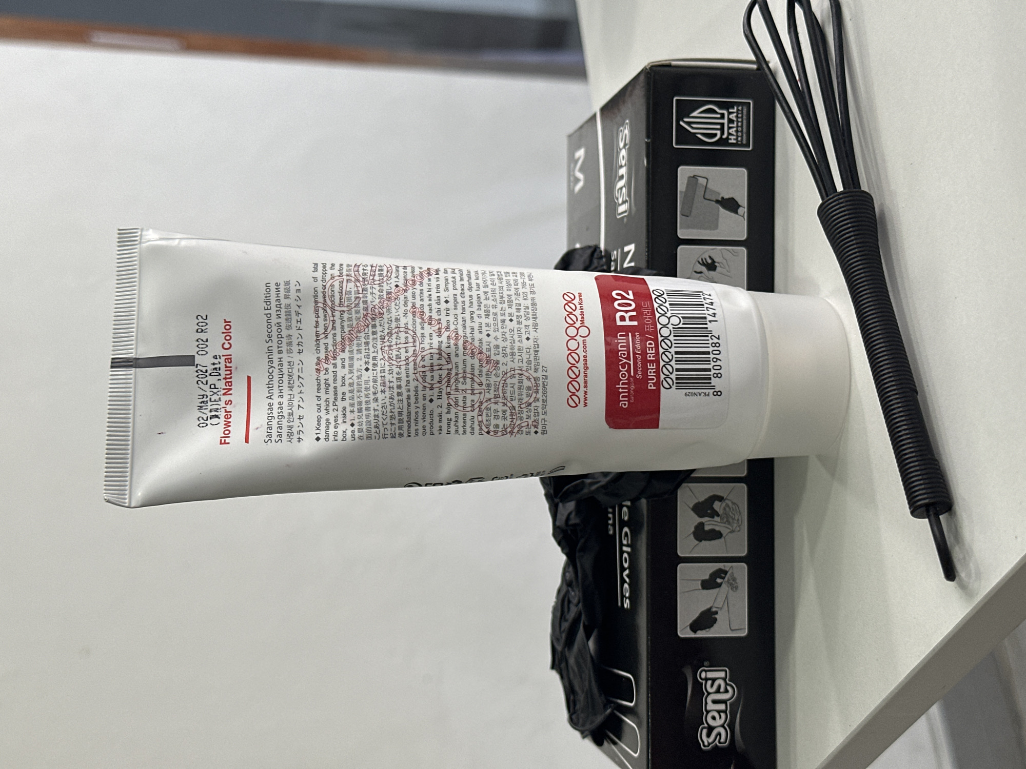 The image shows a white tube of hair dye, possibly Sarangsae Anthocyanin Second Edition, with the color Pure Red indicated on the label. Next to the tube are black gloves in a box and a small black whisk. The background is blurred and appears to be an indoor setting, possibly a store or salon. There are no people or any discernible interaction visible in the image. The location of North Kuta, Indonesia is not depicted in the image.