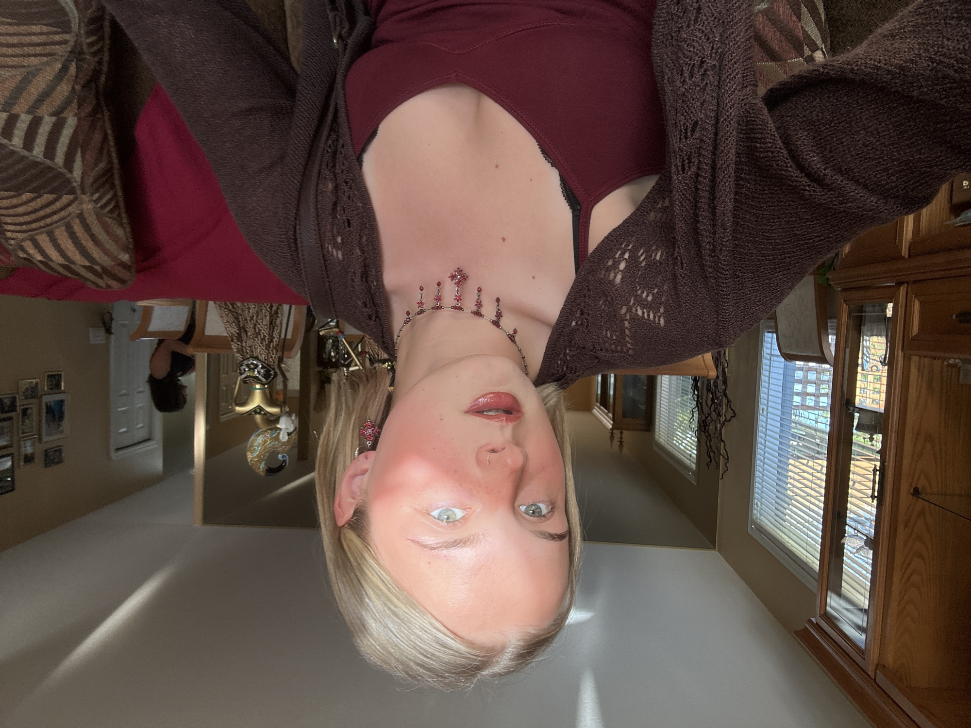 An indoor portrait captures a young person with blonde hair and light-colored eyes, looking directly at the camera with a neutral expression. The individual is wearing a dark red or burgundy camisole under a dark brown, possibly knitted or crocheted, cardigan. A prominent necklace with red jewels, including a central cross-like design, adorns their neck, complemented by matching red jeweled earrings.
The setting appears to be a home, possibly in Kelowna, Canada. Natural light streams through windows with horizontal blinds in the background, casting shadows on the floor, indicating daytime. To the left, part of a patterned cushion or sofa is visible. To the right, a wooden cabinet with glass doors is prominent. Further back, a wall displays multiple framed pictures, and a mirror reflects an adjacent hallway or room. Decorative elements, such as a gold ornament and dried plant arrangements, are scattered on furniture. No visible text is present.