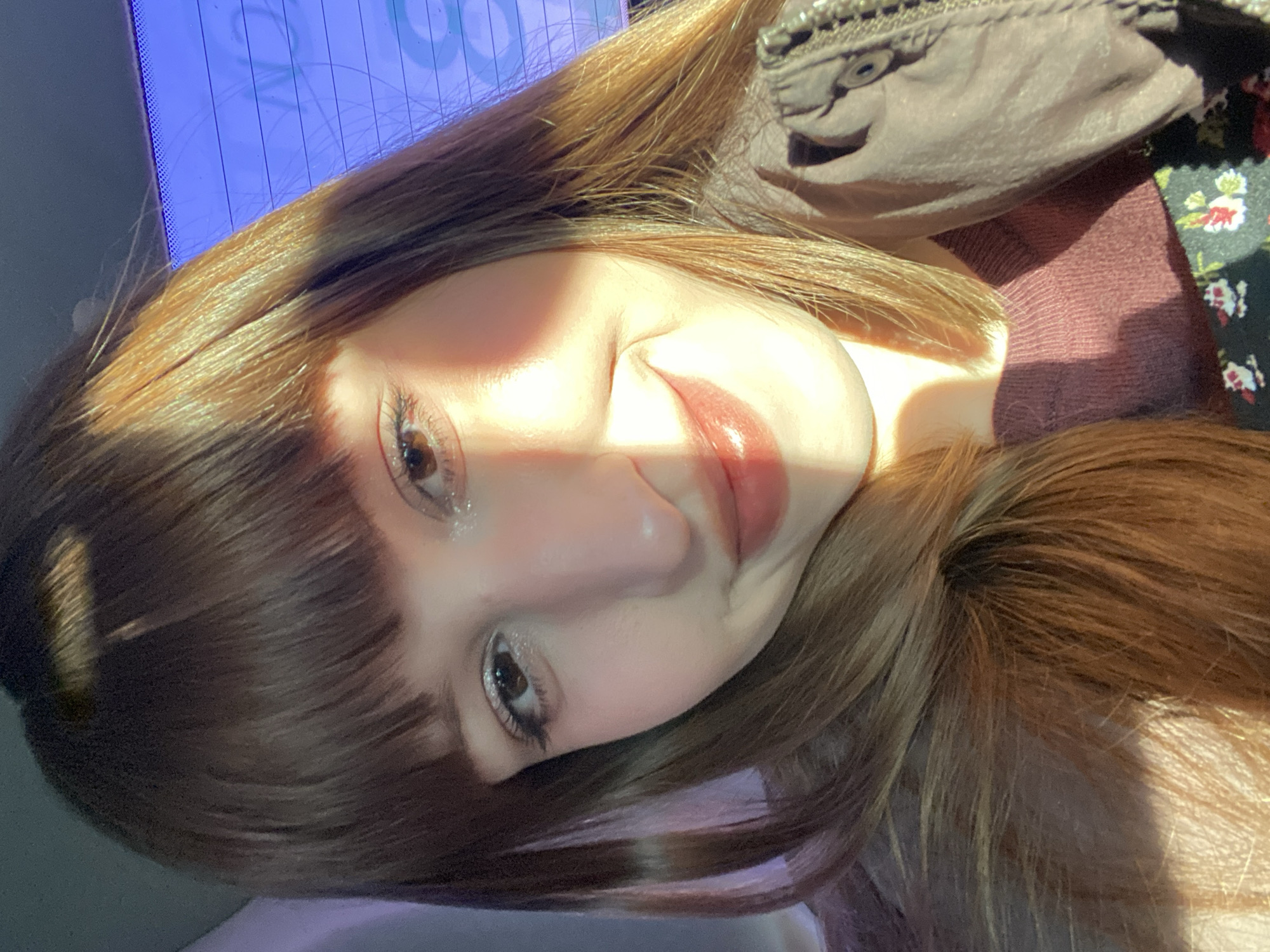 A close-up, portrait-style photograph depicts a young woman with light skin and long, brown hair with bangs. She is turned at an angle to the camera, with a portion of her face obscured by a bright beam of sunlight. Her eyes are visible and appear to be brown. She wears makeup, including eyeshadow and lipstick.
The woman is wearing a brown top and a floral print garment is partially visible at the right side of the frame. A brown jacket or garment is also visible at the top right of the frame, appearing to have some ornamentation.
In the background, a blue surface is partially visible.
The location cannot be definitively determined as Chaikovsky, Russia, from the given image alone. There are no specific landmarks or details that would confirm the setting.