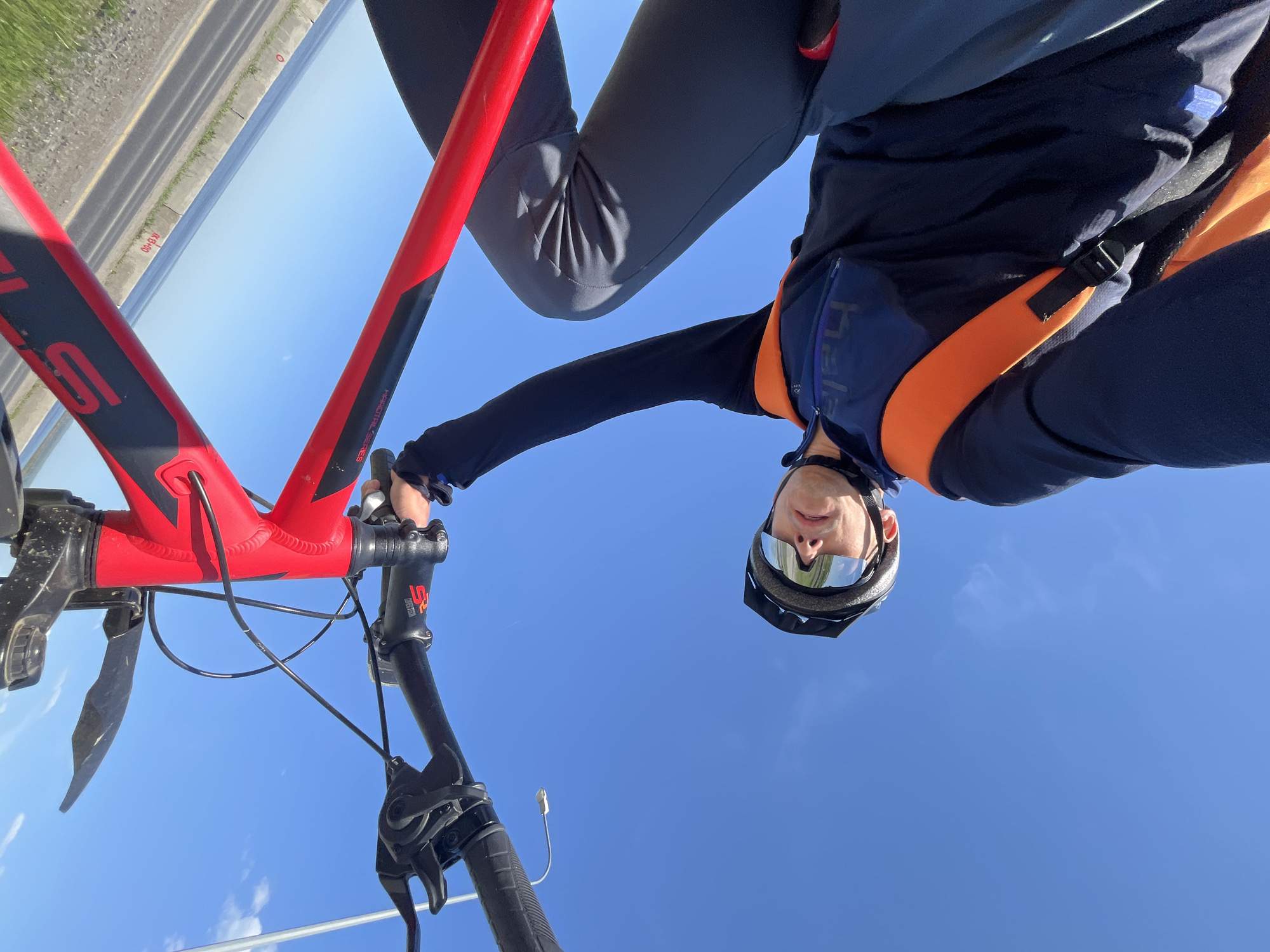 A low-angle shot shows a person on a red and black bicycle against a bright blue sky. The person wears cycling apparel including a helmet and reflective glasses. Part of a road or bridge is visible in the upper left corner. The location is purportedly Chaikovsky, Russia, but this cannot be verified from the image alone.