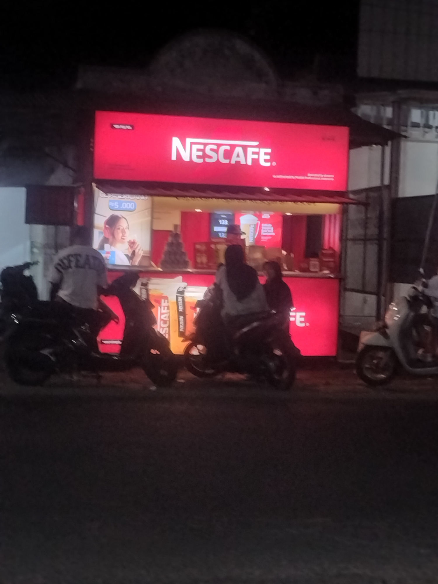 The image captures a nighttime scene outside a Nescafe stand in Srono, Indonesia. A prominent red Nescafe sign is illuminated above a counter where two individuals are visible. One person, wearing a dark hijab, is standing, while another person in a light-colored hoodie is seated on a motorcycle. To the left, a person in a grey t-shirt with the word "NESCAFE" on the back is also seated on a motorcycle, facing the stand. Several motorcycles are parked in front of the stand. The environment appears to be an outdoor urban setting, with a paved surface in the foreground and buildings visible in the background. A white scooter is parked to the right of the scene. The lighting suggests it is after dark, with artificial lights from the stand illuminating the immediate area.
