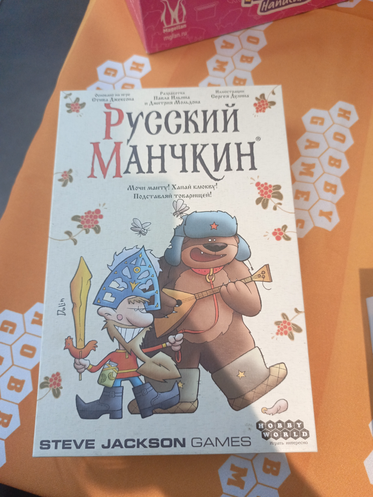 The image shows a board game box for "Russian Munchkin" by Steve Jackson Games. The box is predominantly white with illustrations of a cartoonish character resembling a Russian peasant with a strange helmet and sword, and a cartoonish bear wearing a Russian ushanka hat and playing a balalaika. The text on the box includes information in Russian, such as the game's title, a tagline ("Мочи манту! Хапай клюкву! Подставляй товарищей!"), the names of the game's designers and illustrator, and the tagline for Hobby World, the game's publisher. The box is placed on a table with a patterned orange tablecloth. Other items, like the corner of a box with "Magellan mglan.ru" written on it, are visible in the background. The image doesn't indicate a location in Moscow, Russia, and primarily showcases the board game itself.