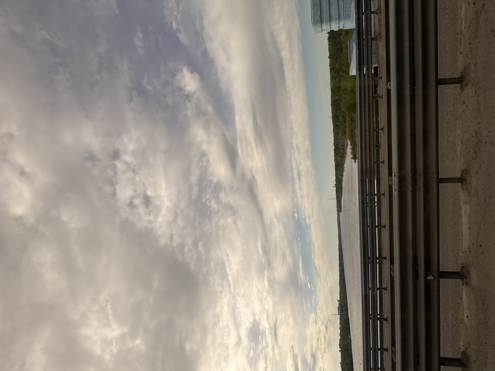 The image shows a view from a bridge in Rusanovo, Russia. A body of water is visible, with a strip of trees on the opposite bank. The sky is filled with clouds, ranging from light to dark gray. The bridge's railing and a section of the bridge structure occupy the foreground. There are no people visible.