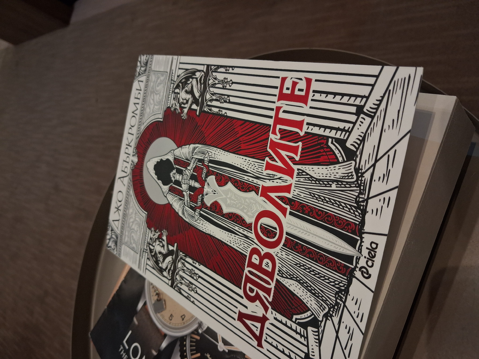 The image shows a book with the title "ДЯВОЛІТЕ" by Джо Аберкромби (Joe Abercrombie), published by @ciela. The book cover features a gothic-style illustration in black, white, and red. A cloaked figure holding a sword is centered, framed by architectural details and gargoyles. The book is placed on top of other items, partially visible, on what appears to be a table or tray. The setting appears to be indoors.