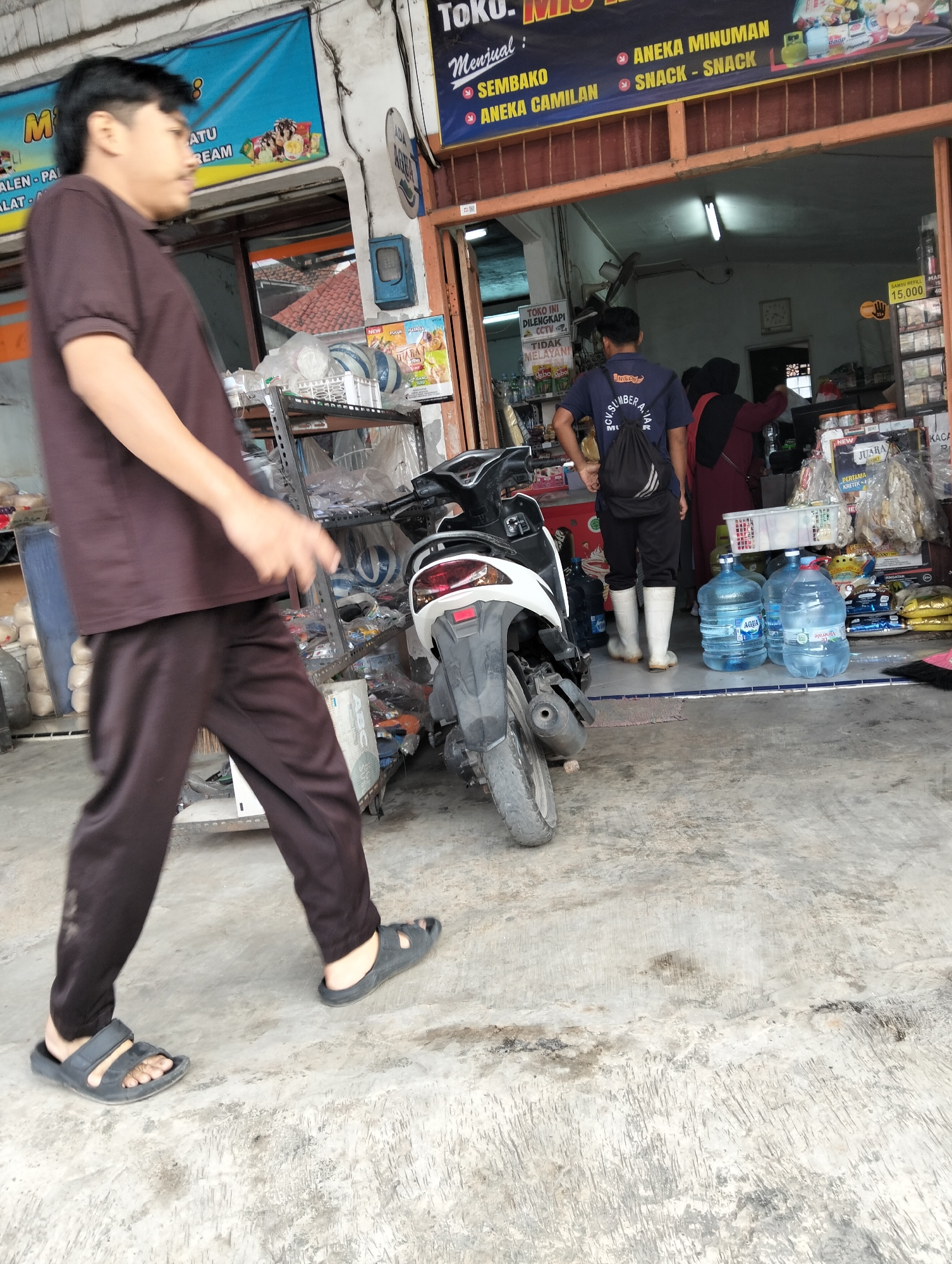 A side view shows a person in dark brown polo shirt and matching pants walking in front of a store in Muncar, Indonesia. The person wears black sandals. A white motorcycle is parked to the right of the person. Several large bottles of water are visible on the ground in front of the store. The store has a blue and yellow sign that reads "TOKO. MUNCAR ANIKA MINUMAN". Inside the store, several people are visible. One person in a blue shirt and black shorts with white boots is facing away from the camera. Another person in a maroon headscarf and matching dress is also inside the store. Shelves are stocked with various items. The ground is concrete.