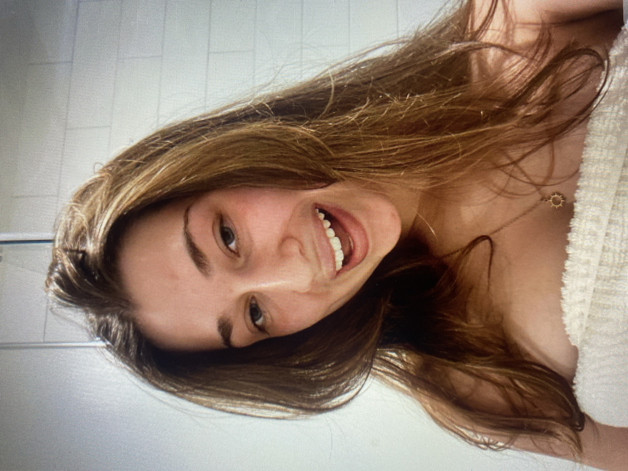 A young woman with long, brown, wavy hair smiles broadly at the camera. She is wearing a towel around her chest and a delicate gold necklace with a sun pendant. The background appears to be a bathroom with white tiled walls. The image is a selfie, taken from a slightly high angle.