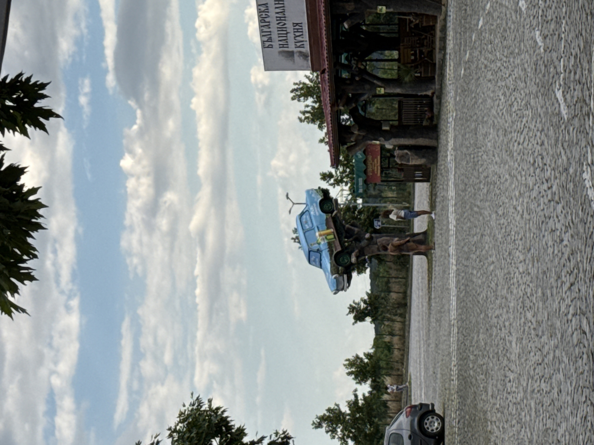 The image depicts an outdoor scene in Polsko Padarevo, Bulgaria, featuring a unique roadside attraction. A light blue car is perched precariously on a stylized tree structure, seemingly as a decorative element. The car is positioned at an angle, with its rear end higher than its front. It has several items visible inside, including what appear to be bottles.
Beneath the car and its tree support, there is a covered outdoor seating area with wooden pillars designed to resemble tree trunks. A sign with Bulgarian text, reading "БЪЛГАРСКА НАЦИОНАЛНА КУХНЯ" (Bulgarian National Cuisine), is visible above the structure.
In the foreground, a cobblestone paved area suggests a parking lot or walkway. A dark grey car is parked on the left side of the frame. Several individuals are present. A woman in a white shirt and blue shorts stands near the car on the tree structure. Another person, appearing to be a man with brown skin and wearing a brown garment, stands near the base of the tree support. Further in the background, a woman in a white t-shirt and denim shorts is visible, and another person is seen walking near a fence line.
The environment is characterized by greenery, with trees and shrubs lining the background. The sky is partly cloudy, with white cumulus clouds scattered across a pale blue expanse. The overall impression is a roadside establishment that uses an unusual display to attract attention.