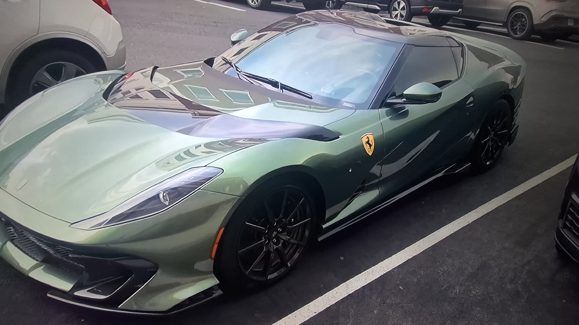 The image captures a green Ferrari 812 Competizione parked in an outdoor parking lot. The car is positioned diagonally in a parking space marked by white lines. A silver-colored vehicle is parked adjacent to the Ferrari on the left. In the background, multiple cars are parked, and the architecture of a building is visible in the reflections on the car's windshield. The environment suggests an urban setting. The presence of the Ferrari, a high-performance sports car, is the central element, drawing attention due to its distinct color and design.