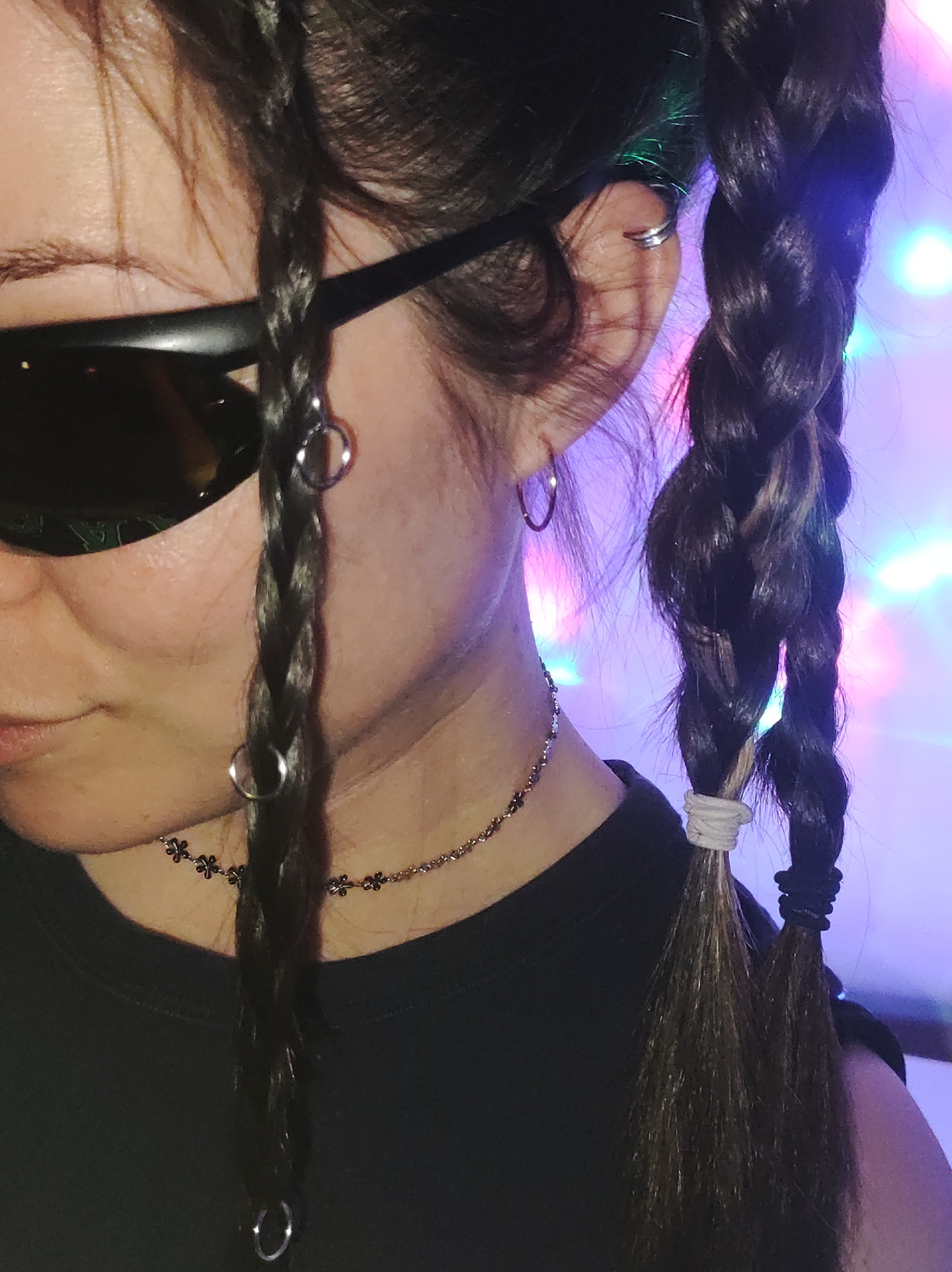 This close-up image captures a person, likely female, from the left side of their head and upper torso. The individual wears dark sunglasses that obscure most of their eyes and nose. Their dark hair is styled in multiple braids; a thinner braid hangs near the face, decorated with two small silver rings. A thicker braid is visible further back, secured with a light-colored elastic, with another braid tied with a dark elastic. The person's left ear is adorned with a small silver hoop earring in the lobe and a silver cuff on the helix. Around their neck is a delicate chain necklace featuring small flower-shaped charms. They are wearing a dark-colored top.
The background is softly blurred with colorful lights, predominantly pink, purple, and blue, suggesting an indoor setting with ambient or party lighting, likely at night or in the evening. The person appears to be posing for the photograph. There is no visible text in the image. While the location context of "Фоки, Russia" is provided, no direct visual cues within the image confirm this specific location.