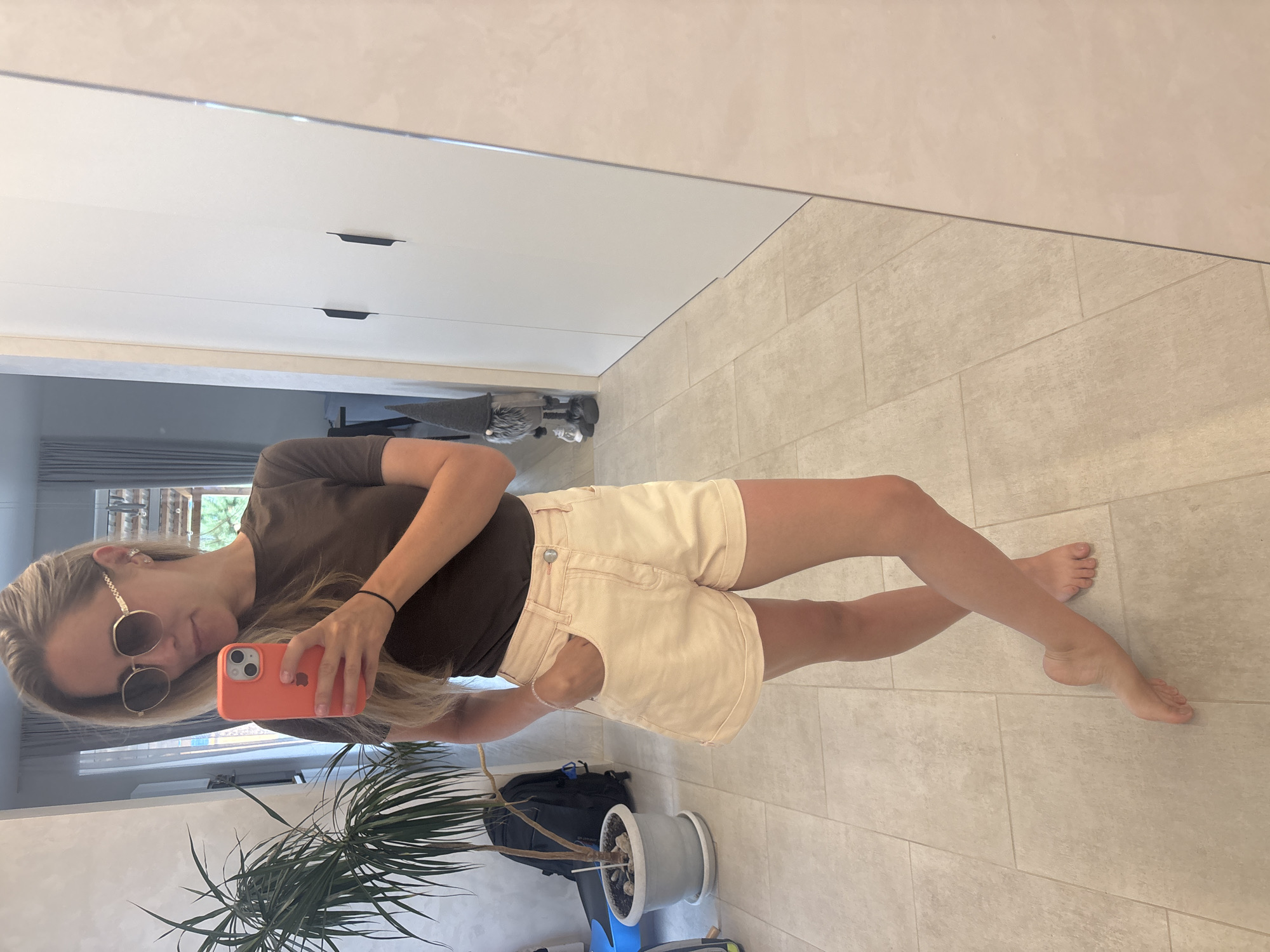 A blonde woman is taking a selfie in a bright, modern interior space, likely a home. She is wearing a dark brown t-shirt and light peach-colored denim shorts. Gold-rimmed sunglasses with brown lenses obscure her eyes, and she is holding a coral-colored smartphone in her right hand, angled to capture her reflection. Her left hand is in the pocket of her shorts. Her legs are crossed at the ankles, and her bare feet are positioned on a light-colored tiled floor.
Behind her, to the left, is a tall potted plant with long, slender leaves, and a black backpack leaning against it. Further in the background, a gray curtain is visible, and a small gnome figurine with a gray hat stands near a large white wardrobe with two dark handles. The overall environment appears clean and organized, with neutral-toned walls and flooring. The reflection in the mirror suggests the space is a hallway or entryway. The image quality is clear, allowing for detailed observation of the subjects and their surroundings. The lighting is even and diffused, indicating an indoor setting with possibly natural light from windows not directly visible. There are no indications of this being in Chaikovsky, Russia.