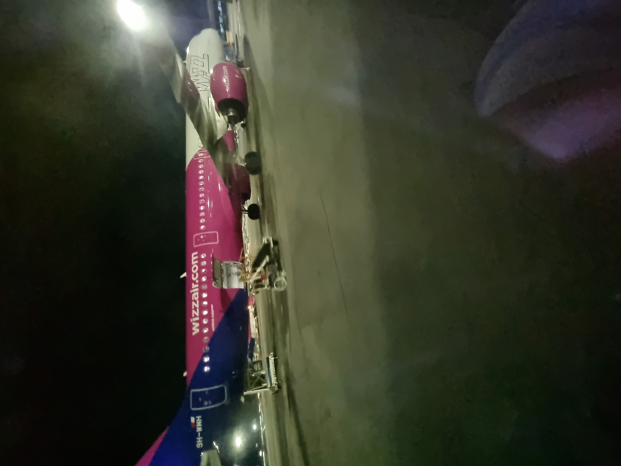 The image shows a Wizz Air Airbus A321 aircraft on the ground, likely at an airport. The fuselage of the plane is predominantly pink with "wizzair.com" printed along the side. The tail is blue with registration number "9H-WMH". The aircraft is parked on a concrete surface, with a boarding ramp or access equipment visible near the fuselage. It is dark, suggesting the photo was taken at night or in low light conditions. Given the registration number and presence of Wizz Air, it is possible that the location is the airport in Sofia, Bulgaria, where Wizz Air operates flights.
