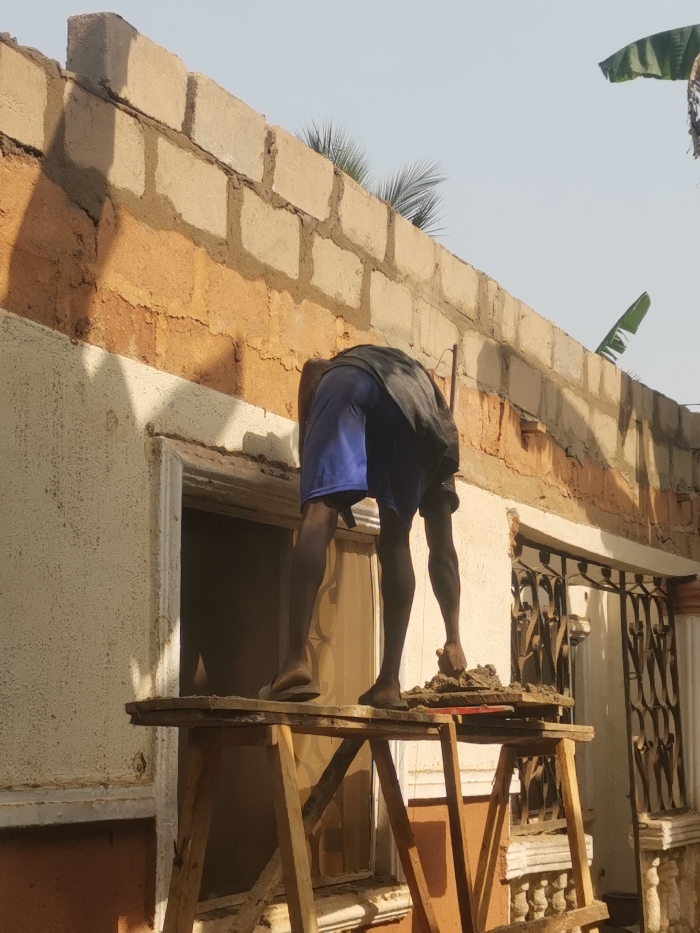 The image depicts an outdoor construction or renovation scene on a building facade in Agbor, Nigeria, under clear daylight. A lone male individual is observed from behind, bent forward over a section of the wall above an open window frame. He is wearing a dark grey t-shirt and blue shorts, with his right foot bare and his left foot wearing a light-colored sandal. The individual stands on a rudimentary wooden scaffold consisting of two A-frame trestles supporting a single plank. A pile of reddish-brown, textured construction material, likely mortar or mud, rests on the plank near his right foot. His right hand is positioned close to this material. The building's exterior features a lower wall section with textured, light beige paint over a reddish-brown substrate. Above this, several courses of grey concrete blocks are laid with mortar, indicating ongoing construction or an unfinished upper section. A window opening is central to the image, lacking glass and revealing a translucent screen or curtain inside. To the right of this opening, an ornate dark metal security grill covers another window, with a decorative concrete balustrade detail below it. Palm fronds and banana leaves are visible above the wall in the background against a bright sky. Strong shadows on the facade suggest direct overhead sunlight.