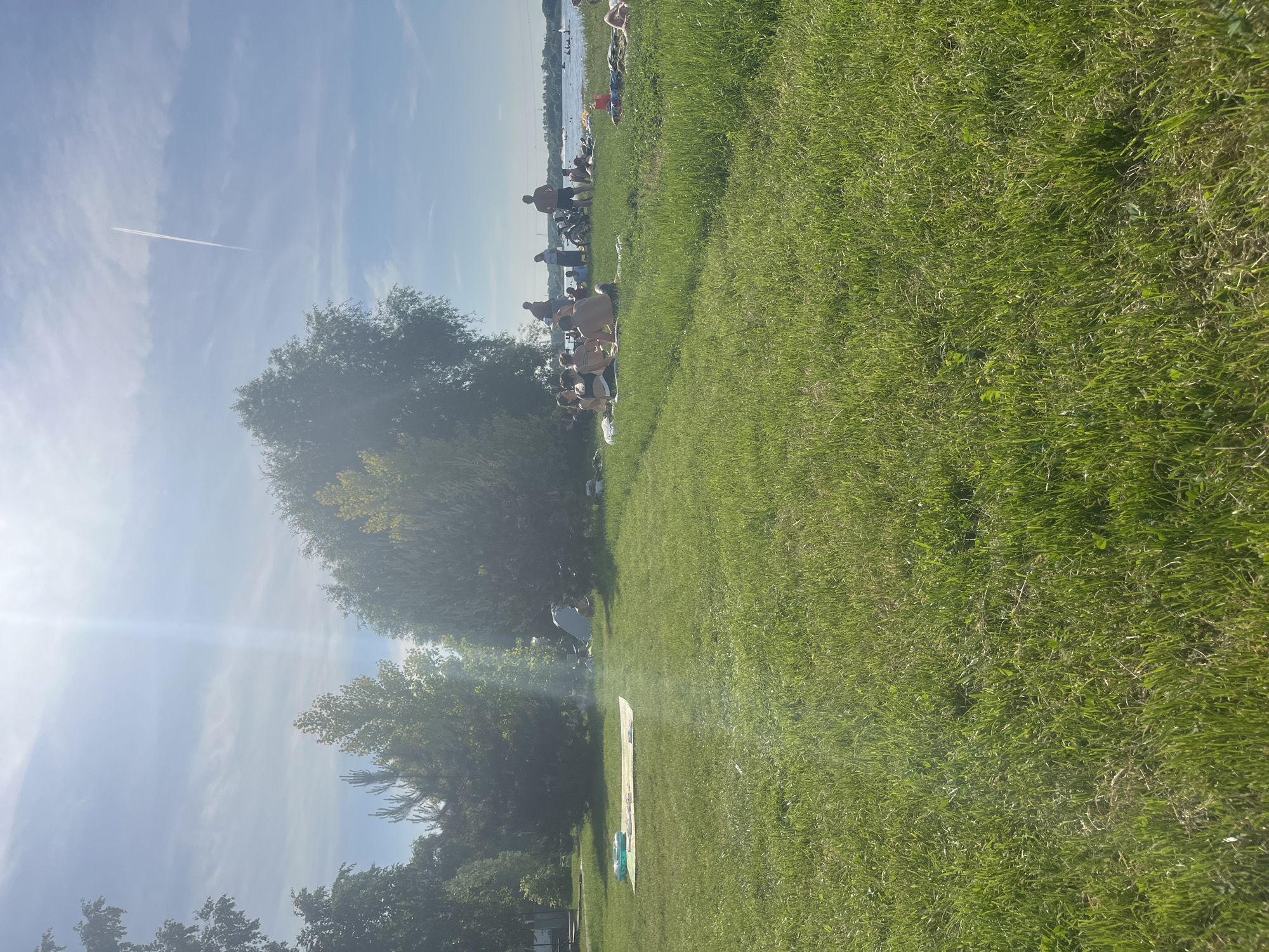 This outdoor recreational scene, situated in Kapustino, Russia, captures a bright, sunny day by a large body of water. The foreground consists of a vast expanse of verdant green grass, showing varying textures and growth patterns. In the left-central foreground, a light-colored rectangular blanket or mat is laid on the grass, topped with a small, light blue inflatable pool.
Further into the midground, a diverse array of deciduous and coniferous trees lines the grassy area, with strong sunlight filtering through their canopies. Behind a cluster of trees on the left, a light-colored tent or temporary shelter is partially visible, with a faint plume of smoke or haze rising nearby.
Closer to the water's edge, predominantly in the right-middle ground, a group of approximately 8-10 individuals is gathered. Most are seated or reclining on the grass, some on blankets or towels, appearing to be sunbathing, socializing, or observing the water. Attire includes swimwear such as bikini tops and shorts, indicating warm weather activities. Two individuals are standing amongst the seated group. A red bucket or similar container is visible near one of the groups on the grass.
Beyond the people, a wide body of water, likely a lake or river, extends to the horizon. The water surface shows slight ripples and reflects the bright sky. In the far distance on the water, several small watercraft or boats are visible, suggesting ongoing recreational activity. The distant shoreline is indistinct, with a low horizon.
The sky in the upper portion of the image is a clear, bright blue with scattered wispy white clouds. A prominent lens flare or sun glare is present in the upper-left quadrant, indicating intense sunlight. A distinct white contrail from an aircraft streaks vertically across the upper-right section of the sky. The overall scene conveys a leisurely outdoor environment on a well-lit day.