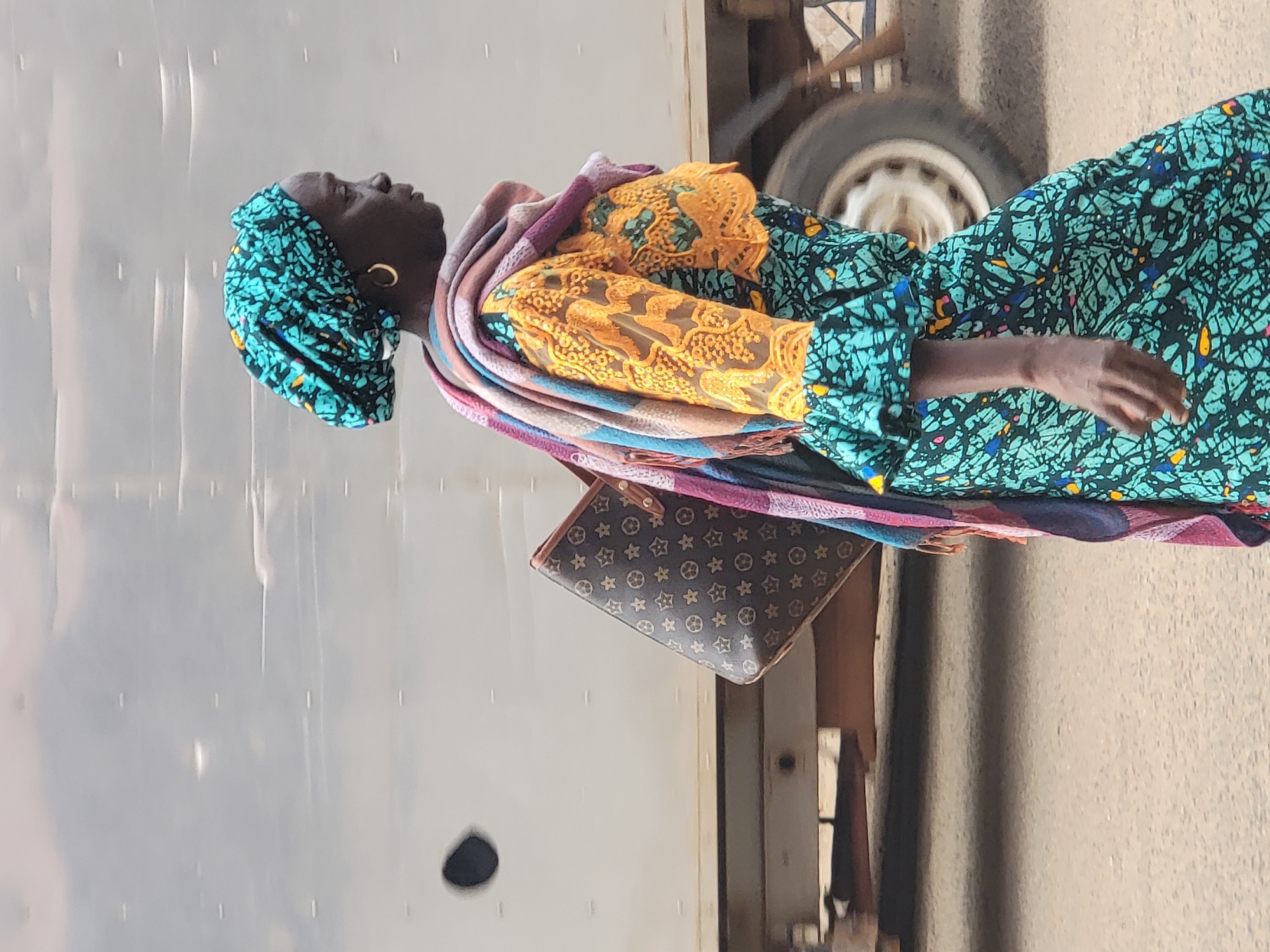 A woman is standing near a building and a vehicle in Maiburiji, Nigeria. She is wearing a patterned teal head wrap, a patterned teal and black long-sleeved dress with yellow lace details, and a colorful scarf draped around her shoulders. She is carrying a brown patterned handbag. She has gold hoop earrings. She is standing on pavement, with a gray corrugated metal wall to her left. To her right, a wheel of a vehicle is visible.