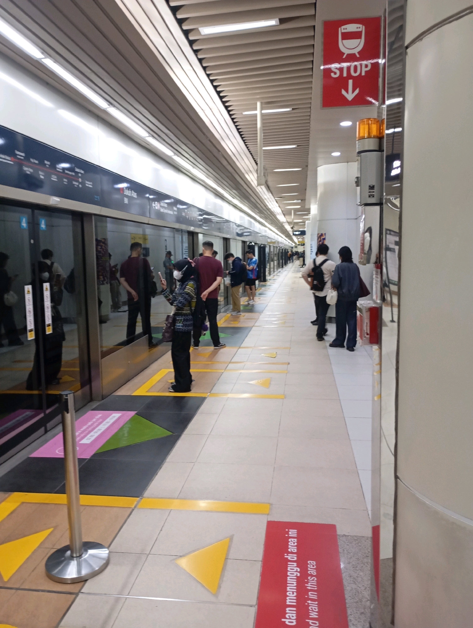 The image shows a platform at a subway station in Central Jakarta, Indonesia. Several people are standing on the platform, waiting for a train. The platform is clean and modern, with yellow and black markings on the floor to indicate waiting areas. A train is visible through the glass doors of the platform screen doors. The lighting is bright and artificial. The overall impression is one of a busy but orderly public transportation hub.