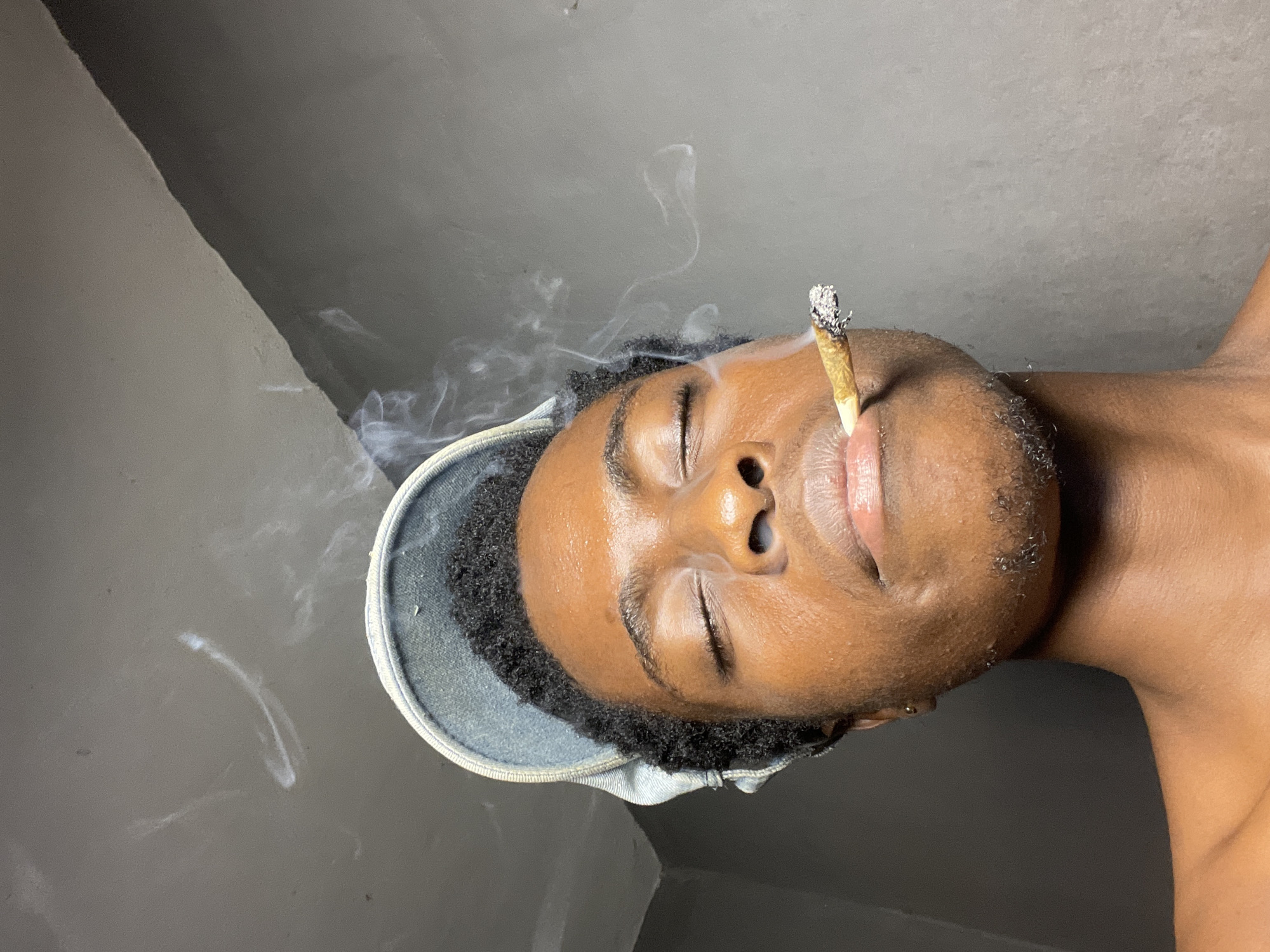 An intimate, close-up, and sideways-oriented shot captures a person with dark skin and short, curly black hair, likely in Nkpor, Nigeria. The individual's eyes are closed, and their mouth is slightly open, holding a lit, hand-rolled cigarette or joint. A distinct plume of white smoke rises from the burning tip, swirling gracefully around their head and against the background. The tip of the joint shows white ash, indicating it is actively burning. The person is wearing a light-colored, possibly denim, cap backward, visible on the left side of the frame. Their skin has a noticeable sheen, reflecting light, suggesting it might be oily or moist. The background consists of plain, dark-grey walls, with subtle variations in shade and a visible corner, indicating an indoor setting. The overall mood conveys relaxation or a state of being lost in thought, possibly induced by the smoking. There is no visible text in the image.