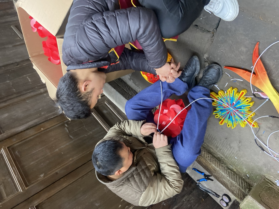 Two men are seated on the floor, likely working on decorations or lanterns. One man wears a black jacket and the other a beige quilted vest. Both are wearing blue jeans. They're surrounded by craft supplies including scissors, pliers, and colorful ornaments made of paper or fabric. One ornament appears to be red flower-shaped and another is yellow with red accents. A cardboard box sits nearby, seemingly containing more craft materials.