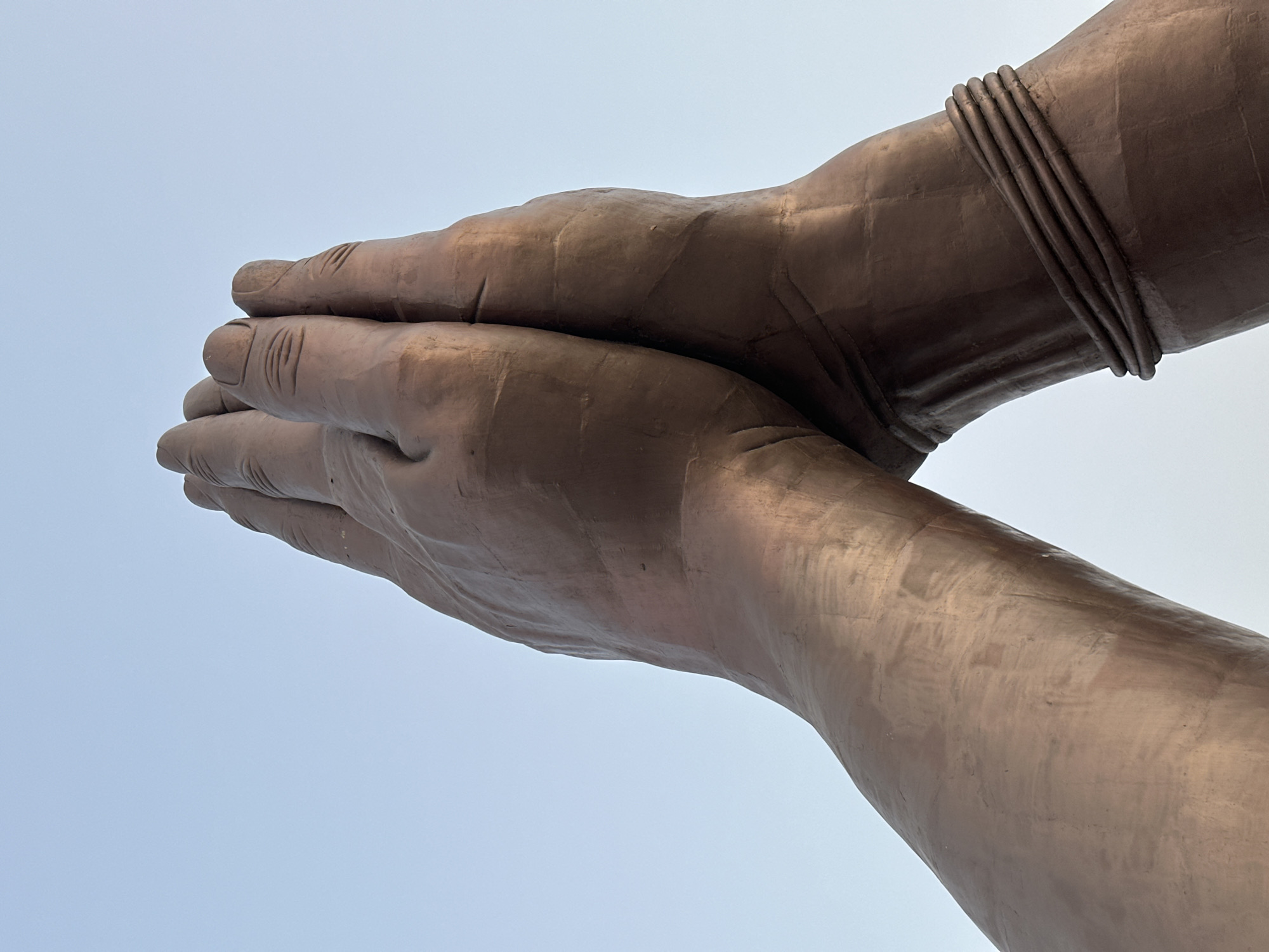 Close-up, upward view of the folded hands of the Statue of Unity in Mughalsarai, India. The hands, made of copper, show detailed texture and are adorned with a bracelet on one wrist. The background is a pale blue sky.