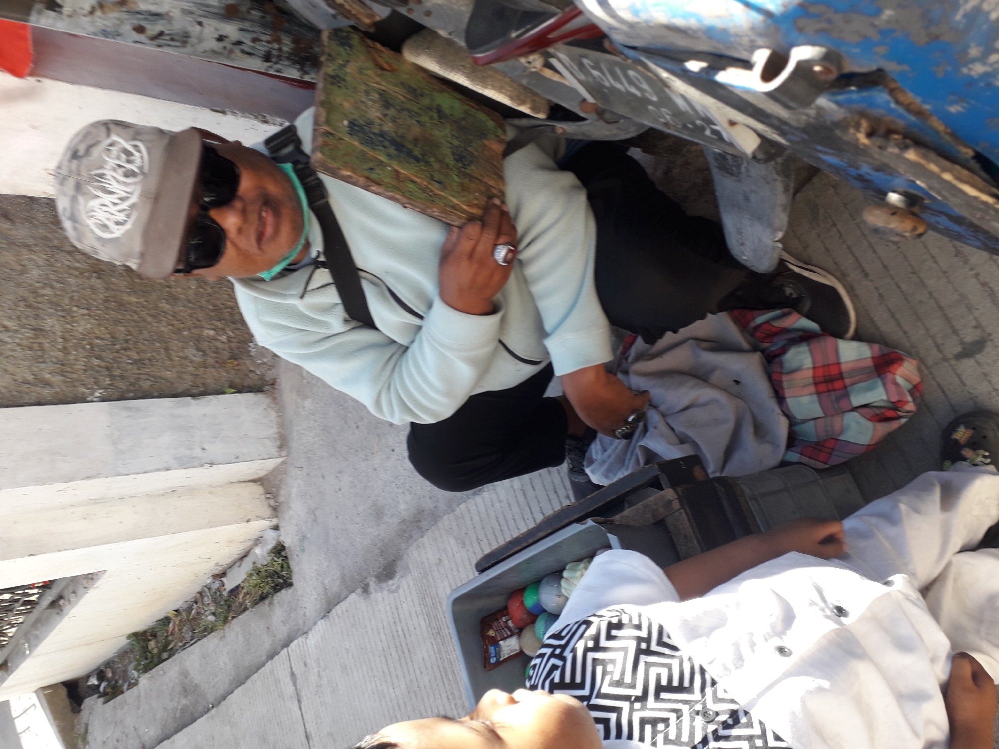 In Cankuang, Indonesia, on a paved surface next to a building, a man wearing a grey baseball cap and sunglasses is sitting. He is holding a rectangular, weathered object, possibly made of wood, in his hands. A black strap is visible across his chest, and a light blue medical mask is partially pulled down around his neck. In front of him is a grey plastic bin containing several colorful balls, and a young child wearing a white shirt with a black geometric pattern is partially visible to the left, looking towards the man. A folded blanket with a red and blue plaid pattern lies on the ground near the man's legs, and a motorcycle is partially visible on the right side of the frame.