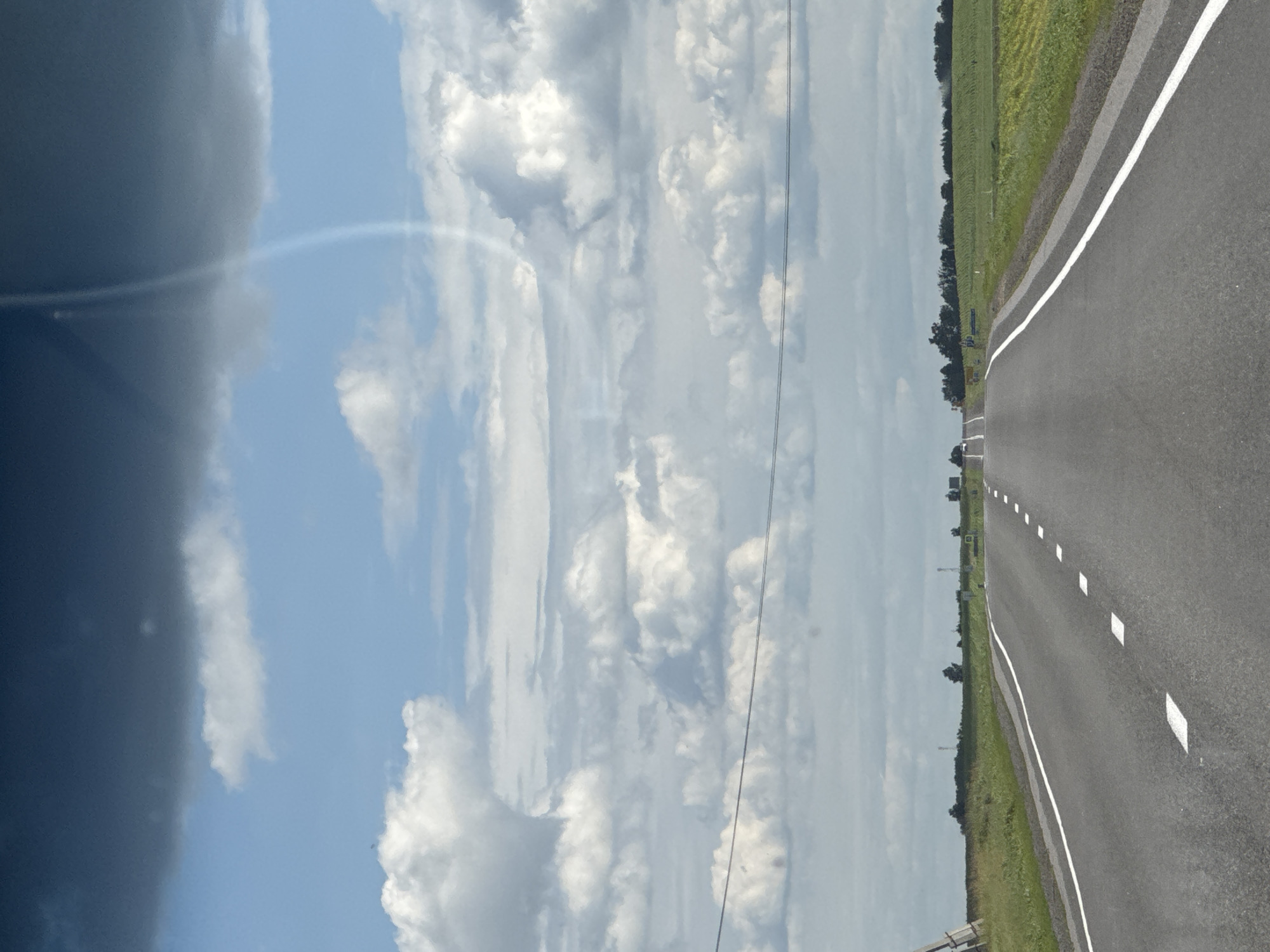 This is an outdoor, eye-level, daylight, wide-angle shot of a paved road in the city of Акцябрскі сельскі Савет, Belarus. The road has white dashed lines on the right side, indicating the edge of the lane, and solid white lines on the left. The road stretches into the distance, curving slightly to the right. On either side of the road are fields of green crops. The sky is filled with large, puffy white clouds and some darker gray clouds. In the far distance, there are trees and what appear to be cell towers. There is a thin, curved streak of light or reflection visible in the upper right portion of the sky. No people are visible in the image.