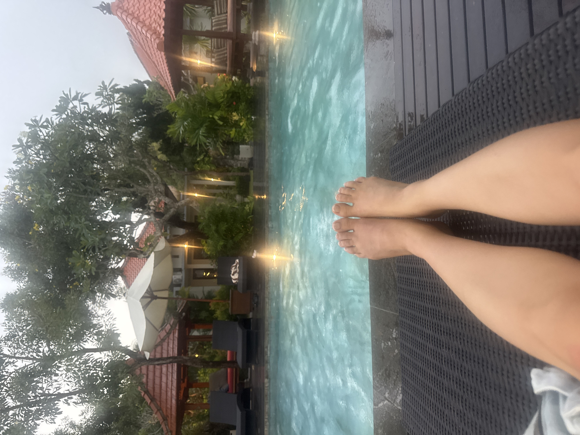 The image shows a pair of legs and feet extended towards a pool in Kuta Selatan, Indonesia. The person is lying on a dark-colored wicker lounge chair. The pool has light-colored water with reflections. Behind the pool are trees, plants, and buildings with red-tiled roofs. There is also an umbrella visible among the trees. The weather appears overcast.