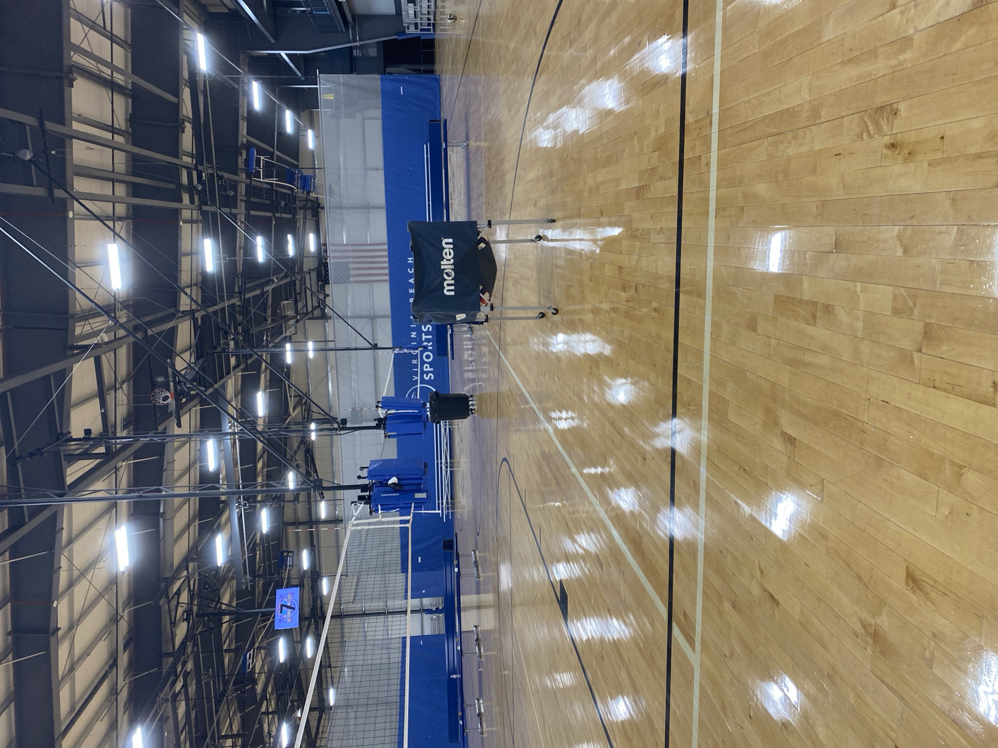 The image shows an indoor volleyball court. The court is made of polished wood flooring with black and white lines marking the boundaries. A volleyball net is set up in the background. There are blue padded protectors on the net poles, and a blue banner with "VIRGINIA BEACH SPORTS" written on it is visible behind the net. A black trash can on wheels is located to the right of the net. In the foreground, a portable ball cart with the "Molten" logo is positioned in the center of the court. The ceiling of the facility features exposed metal beams and industrial lighting. The overall environment appears to be a sports complex or gymnasium.