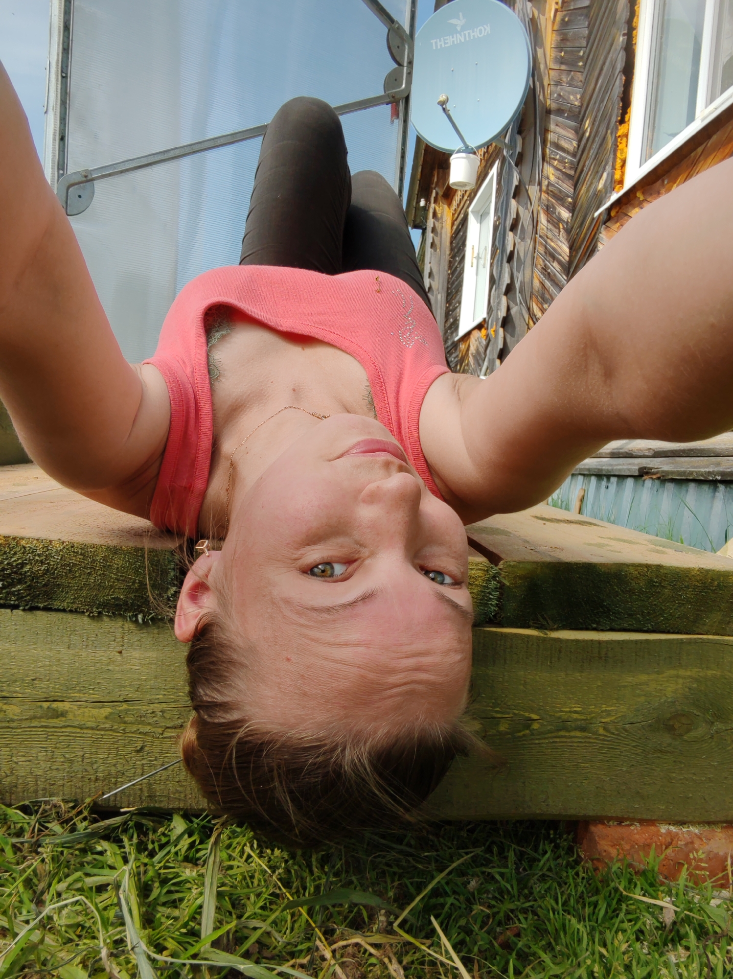A woman is taking a selfie while lying on her back outdoors. She is wearing a pink tank top and dark pants. Behind her, there is a building with a satellite dish and a greenhouse. The background suggests a rural or suburban setting, potentially near a wooden structure. The location is identified as Чухлэм, Russia. There are wooden planks and grass visible beneath her.