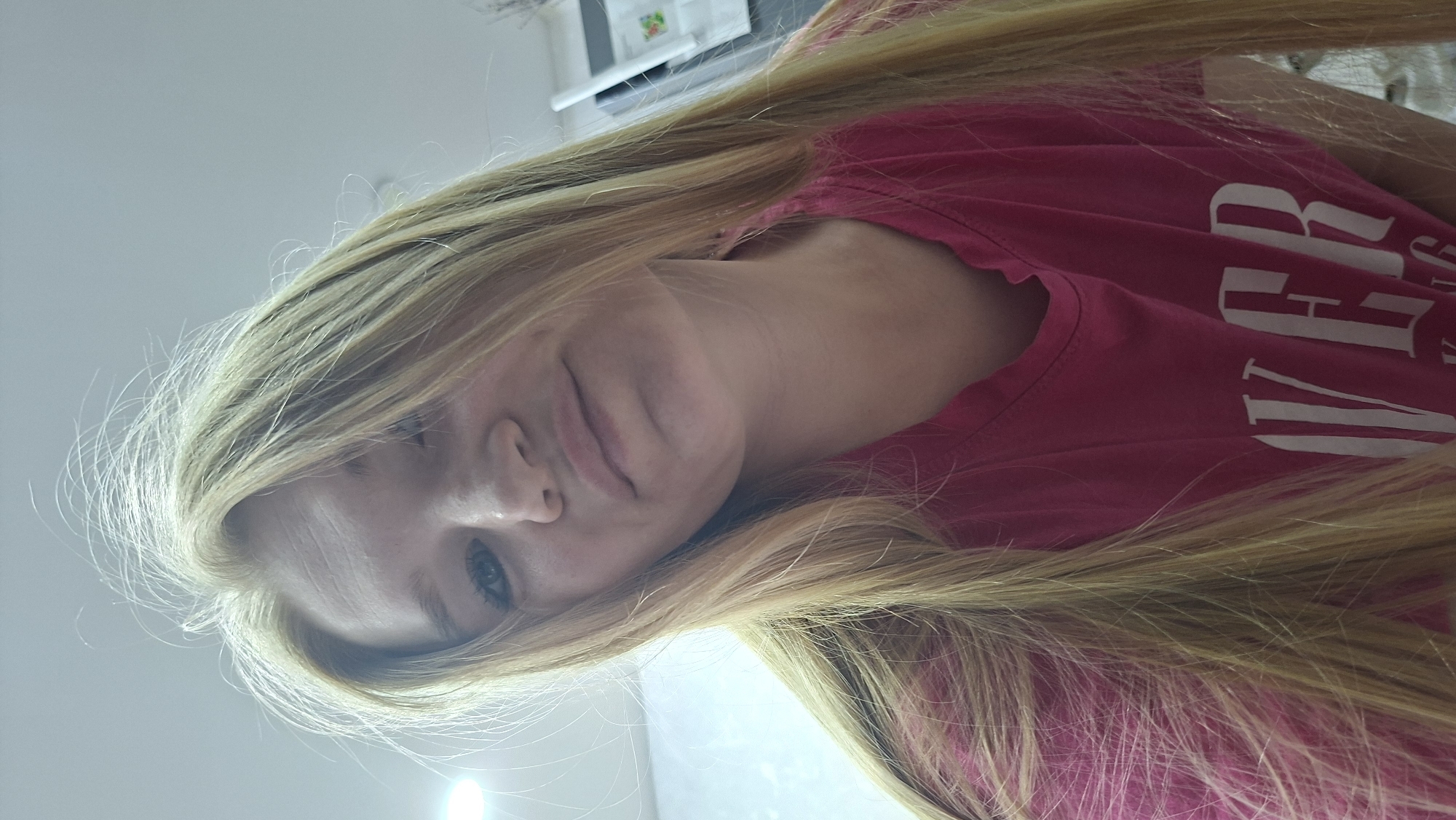The image is a selfie taken by a young Caucasian woman with long blonde hair and blue eyes. She is wearing a pink t-shirt with white lettering, partially visible. The background appears to be an indoor setting with a white wall and a ceiling light. The woman's head is tilted downwards and slightly to the left, and she is looking directly at the camera with a neutral expression. The lighting suggests the photo was taken in natural or artificial indoor light. There is no discernible location in Chaikovsky, Russia.