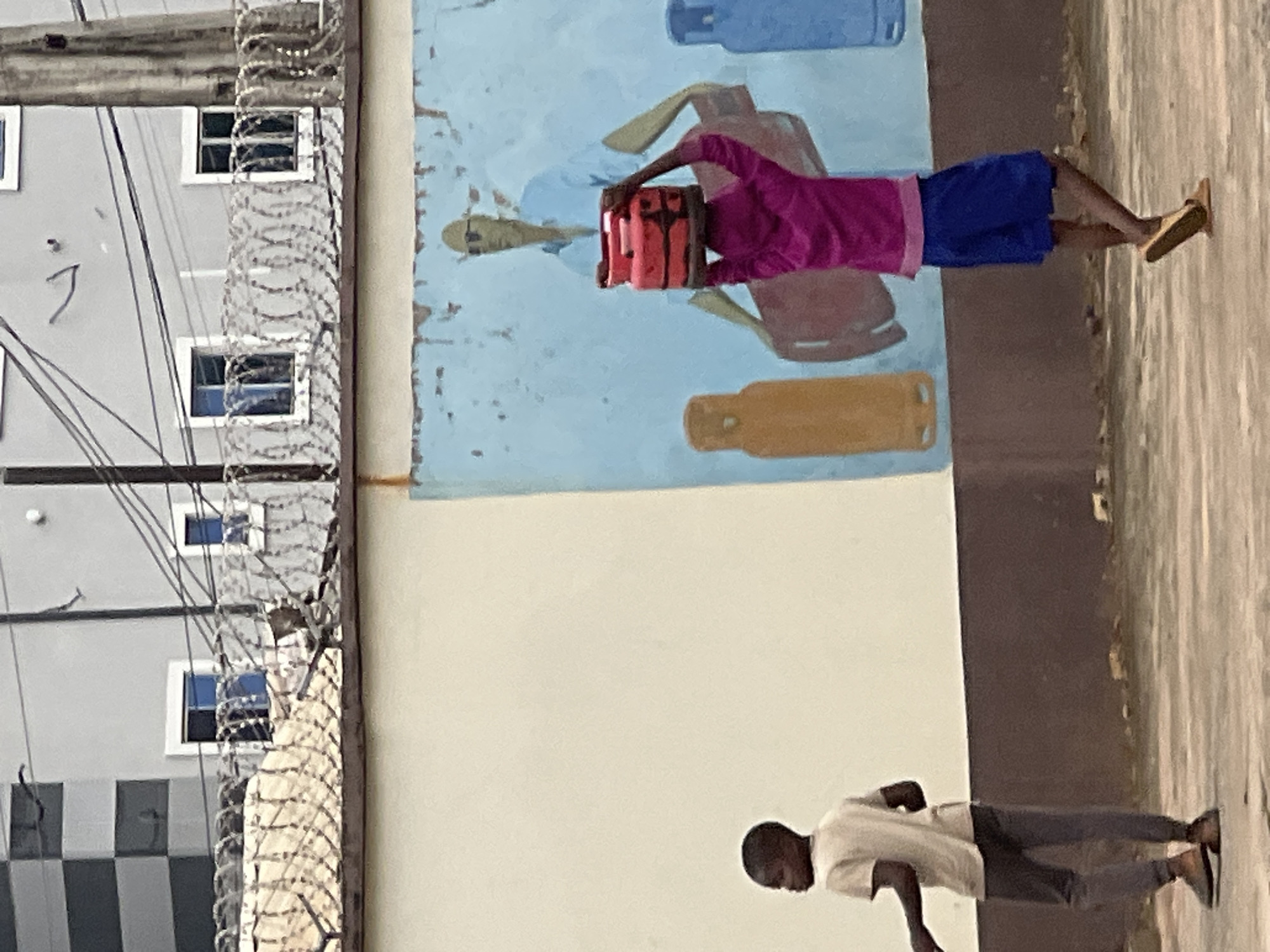 In Nkpor, Nigeria, a young child, possibly a boy, is walking past a mural. The child is wearing a light-colored t-shirt and dark trousers, with brown slip-on shoes. They are facing left, looking away from the viewer. To the right, another child is carrying a stack of red gas cylinders on their head. This child is wearing a bright pink long-sleeved top and blue shorts, with tan slip-on shoes. The mural behind them depicts a bald man in a light blue shirt and sunglasses, and two gas cylinders, one orange and one blue. The scene takes place outdoors, with a weathered wall and barbed wire fence visible in the background, suggesting an urban environment.