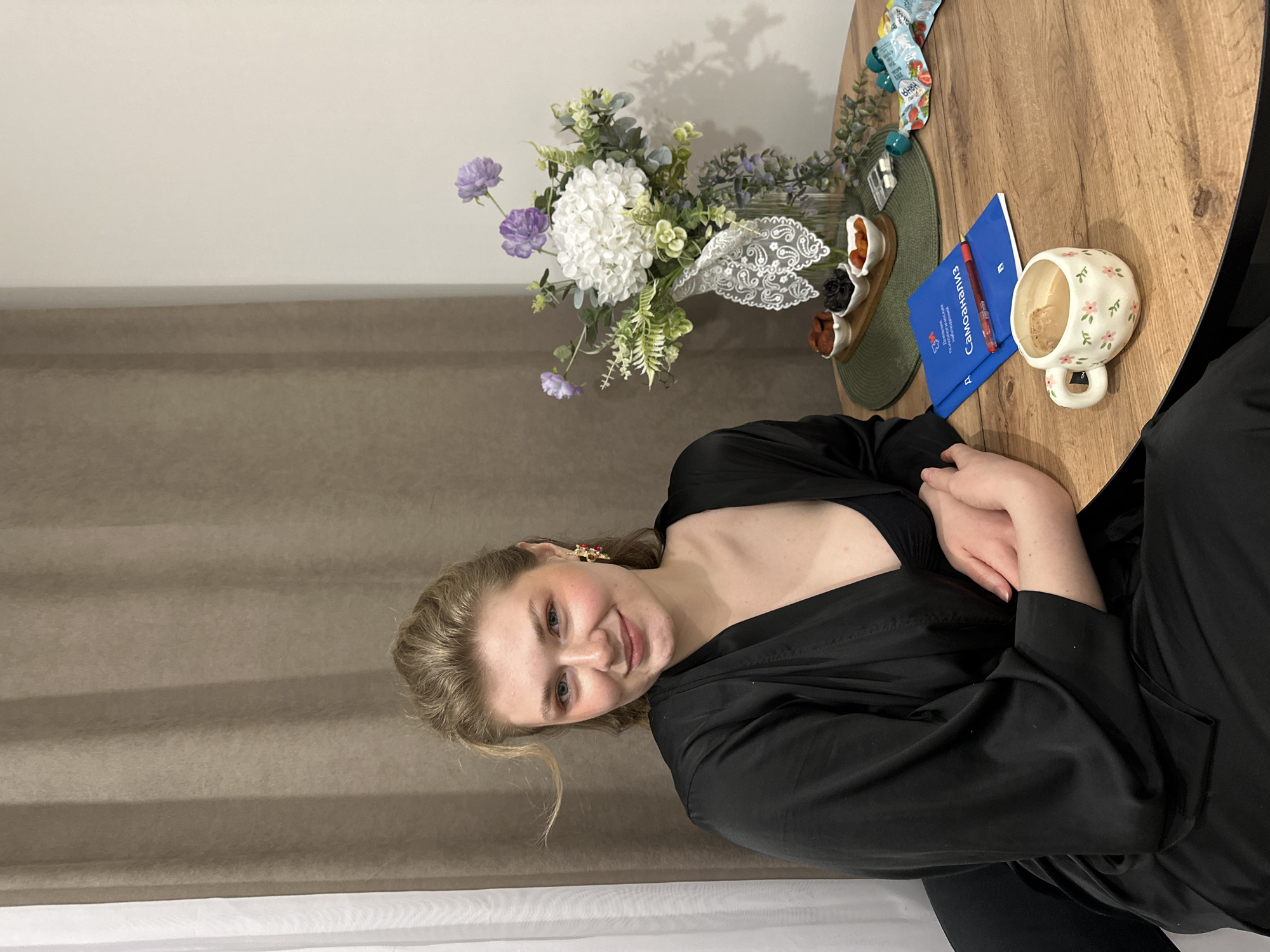 A woman sits at a round wooden table. On the table are flowers in a glass vase, dried fruit in small bowls on a green trivet, a blue document with a red pen, a mug with a flower pattern, and snacks in tubes. The woman is wearing a black robe and smiling. The background consists of a beige curtain and a white wall. This photo appears to be taken in Kazan, Russia.